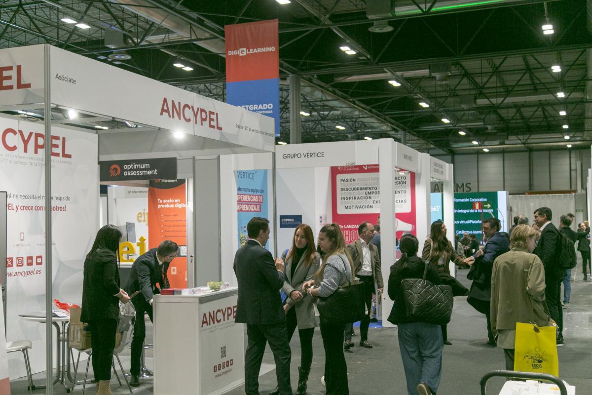 Un pabellón de ferias muy concurrido con numerosos stands de expositores, entre los que destaca uno blanco con el letrero «ANCYPEL» en rojo. Profesionales de negocios, vestidos con traje o con ropa informal de oficina, establecen contactos y conversan.