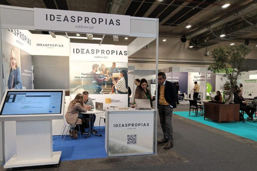 Stand ferial de «Ideaspropias editorial» con visitantes interactuando, una gran pantalla táctil y coloridos carteles. El ambiente es profesional y animado.
