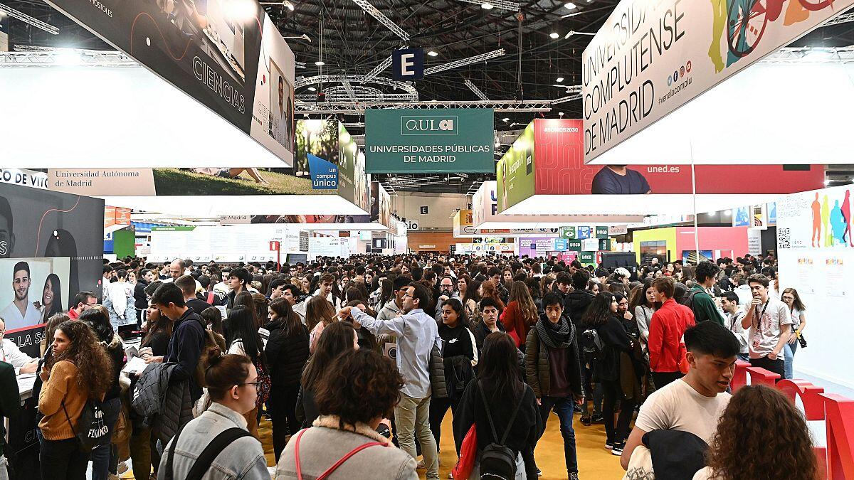 Una gran multitud en una feria educativa cubierta, llena de estudiantes diversos que visitan los distintos stands de las universidades. El ambiente es animado y bullicioso.