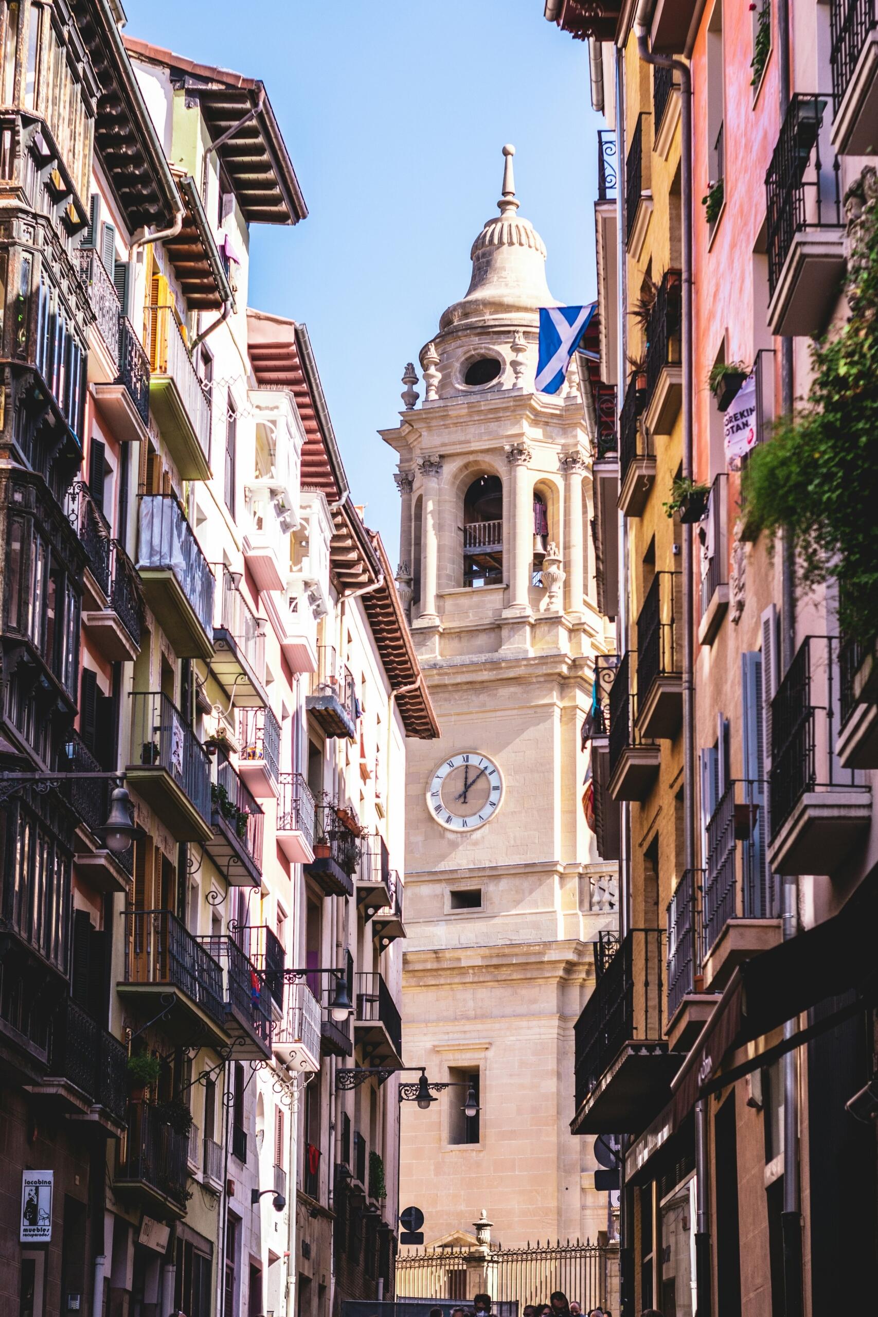 Calle estrecha bordeada de edificios de colores que conduce a una torre del reloj coronada con una cúpula y una bandera escocesa contra un cielo azul despejado en pamplona