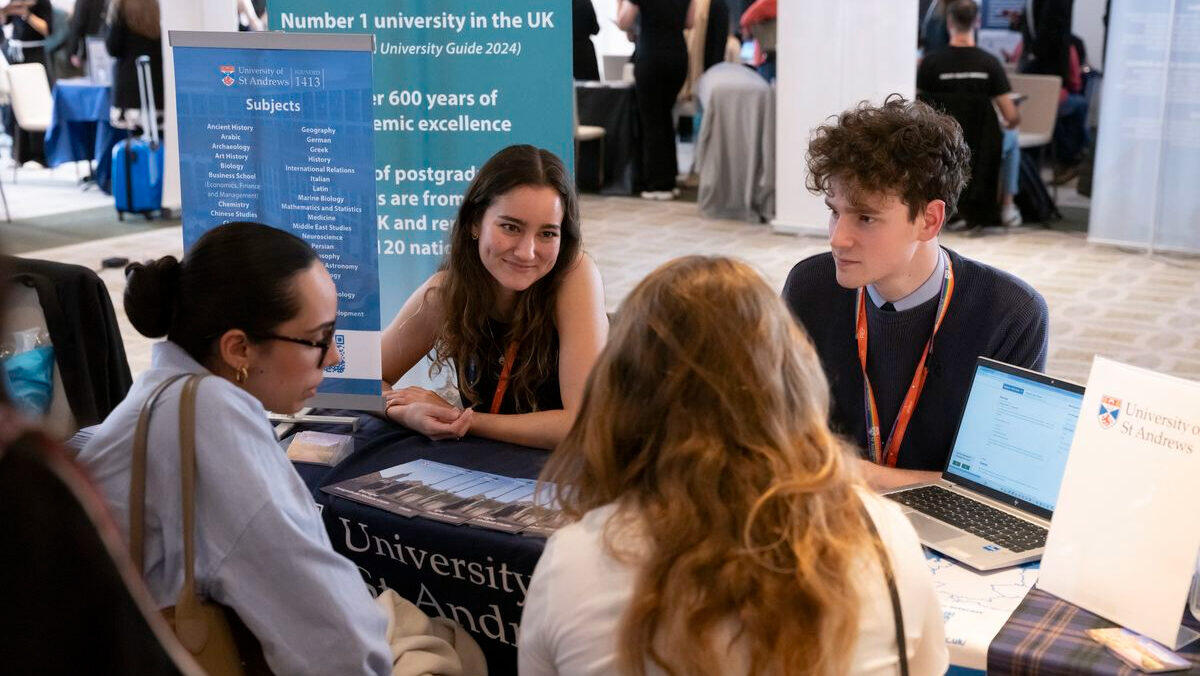 Stand en una feria universitaria con dos representantes interactuando con los estudiantes. Sobre la mesa hay un ordenador portátil y folletos. El ambiente es agradable e informativo.