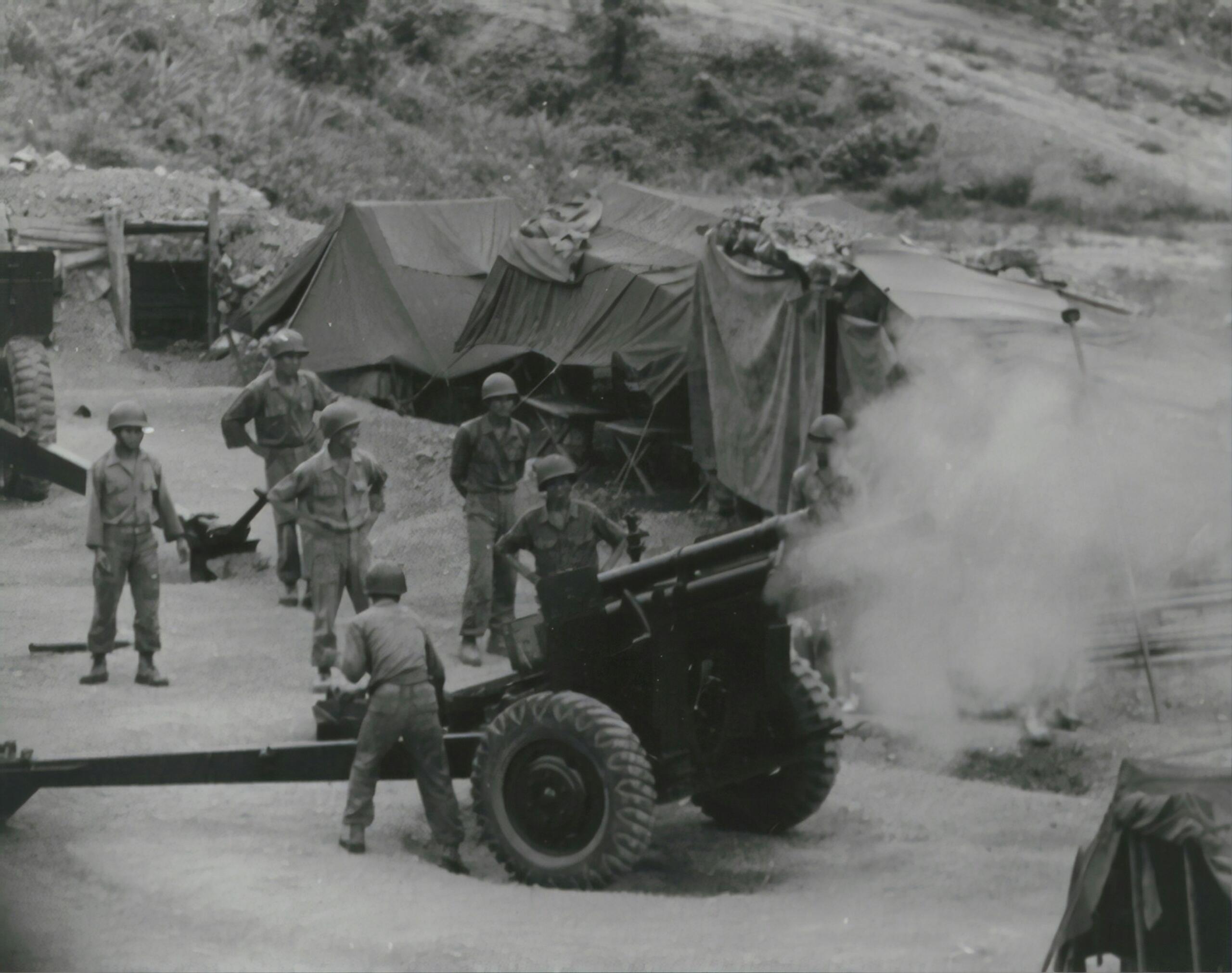 Soldados manejando una pieza de artillería en un campamento militar, con tiendas visibles al fondo y humo saliendo del cañon