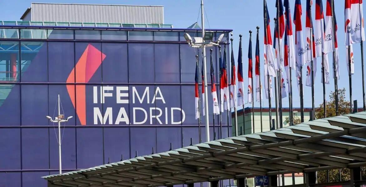 Edificio moderno con las letras «IFEMA MADRID» en negrita sobre la fachada acristalada. Numerosas banderas blancas, rojas y azules se alinean en el lado derecho, lo que sugiere un evento formal.