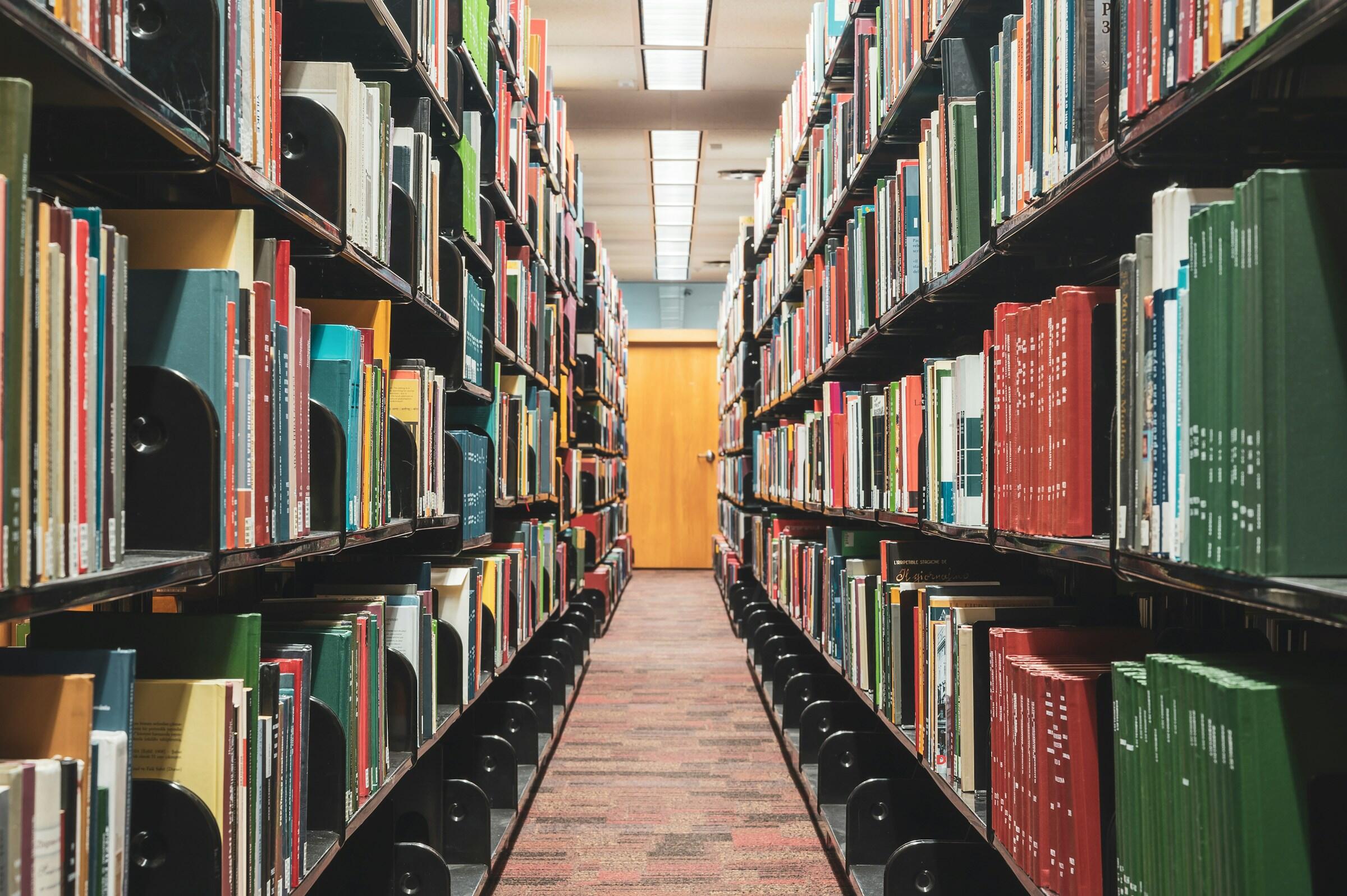 Un largo pasillo entre estanterías altas llenas de libros de colores en una biblioteca, que conduce a una puerta de madera en el extremo más alejado.