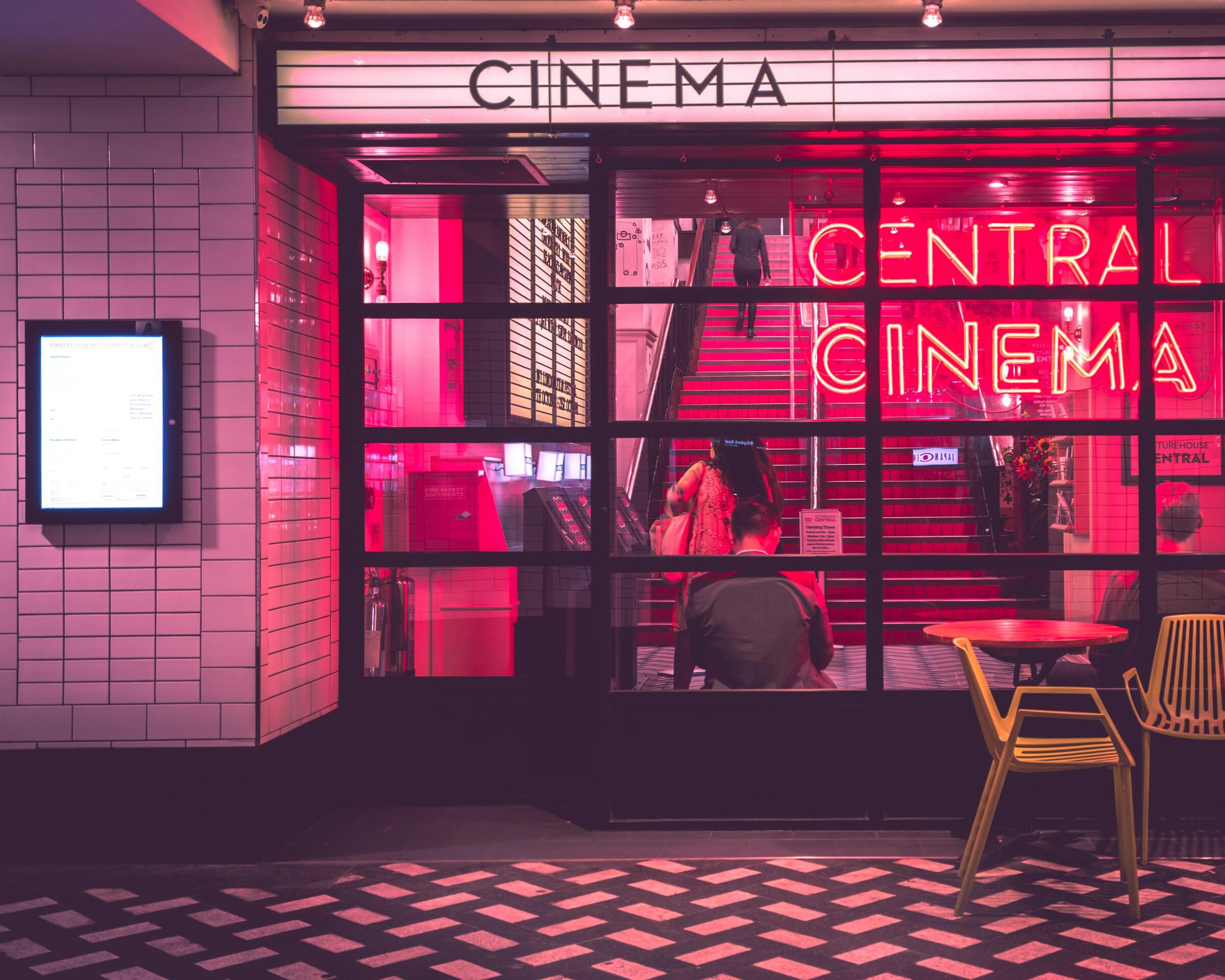 Una vibrante entrada de cine con el letrero de neón «CINE CENTRAL», con una puerta de cristal, un tablón de menús y clientes dirigiéndose al interior.