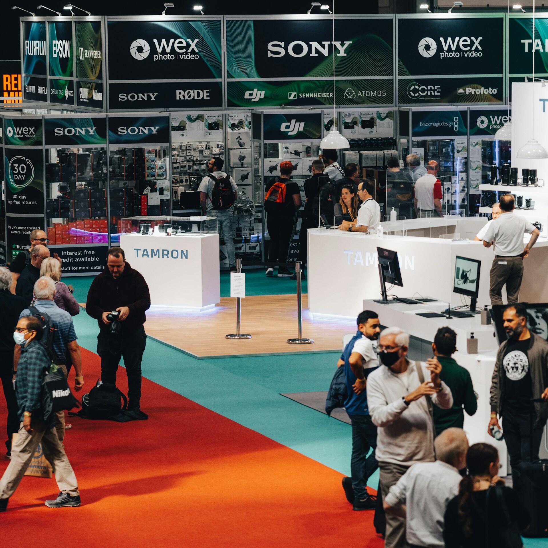 La gente recorre una concurrida feria tecnológica en la que los stands de Tamron y Sony exhiben cámaras y equipos. El ambiente es animado, con alfombras de vivos colores rojo y verde azulado.