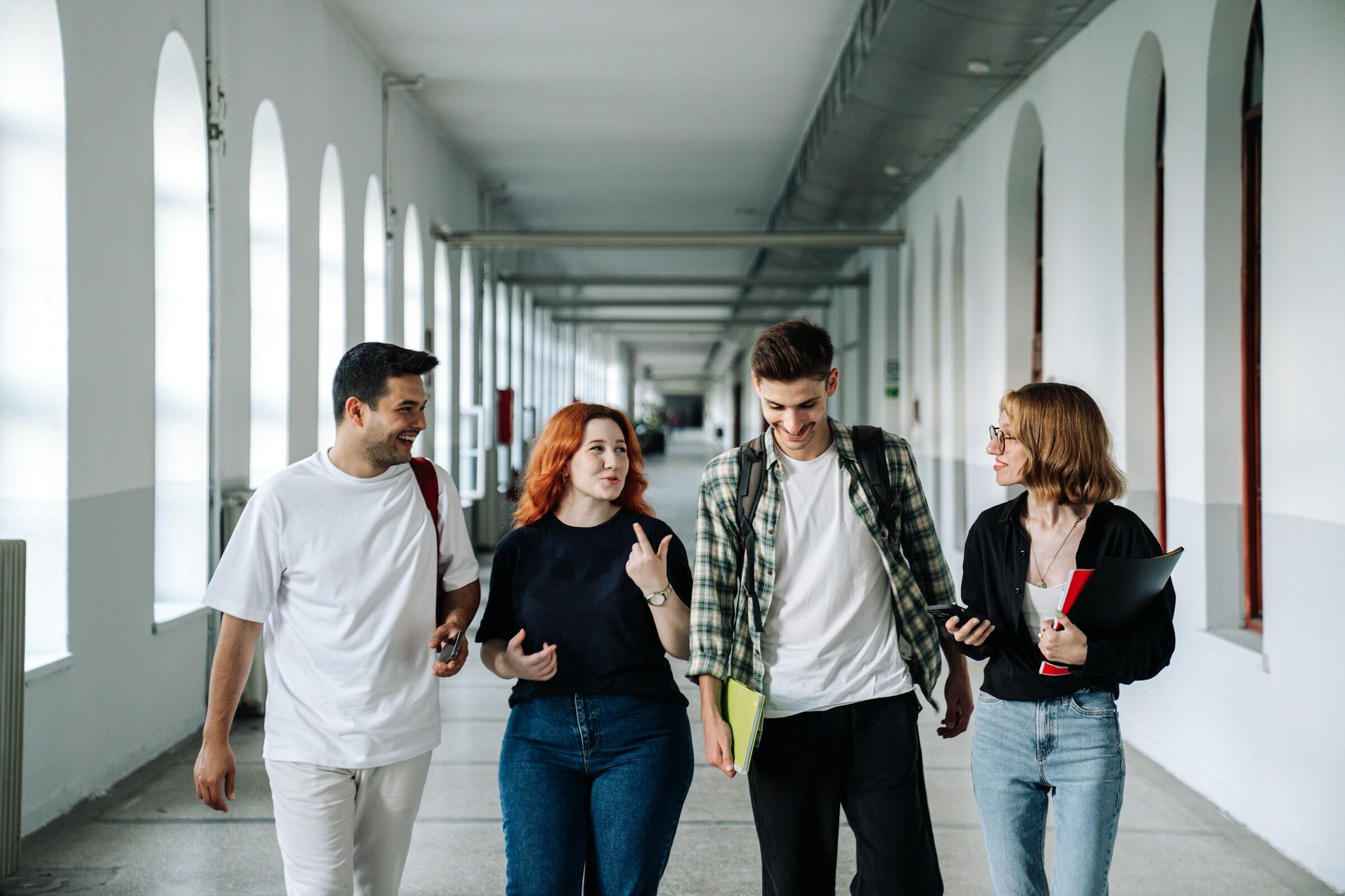 Un grupo de cuatro adultos jóvenes camina despreocupadamente por un luminoso y aireado pasillo bordeado de arcos, enfrascados en una conversación.