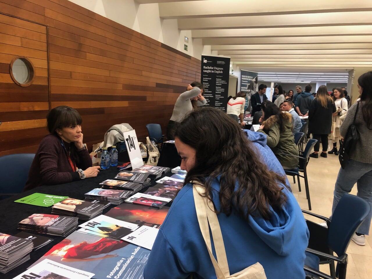 Una concurrida feria de empleo con una mujer sentada en una mesa de información. Hay folletos repartidos y los asistentes conversan con los representantes. El ambiente es animado e informativo.