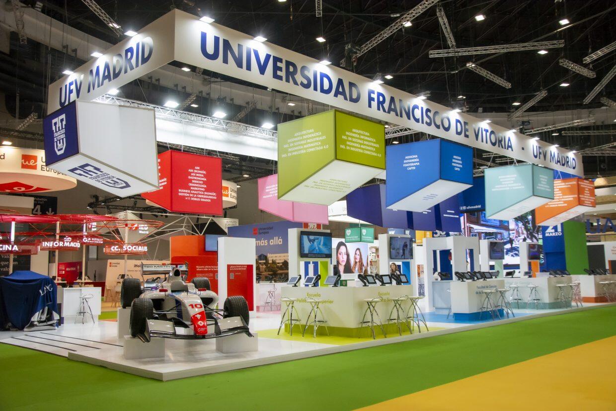 Stand de exposición para la Universidad Francisco de Vitoria, Madrid, con una exhibición de coches de carreras, coloridos carteles colgantes y pantallas interactivas sobre mesas.