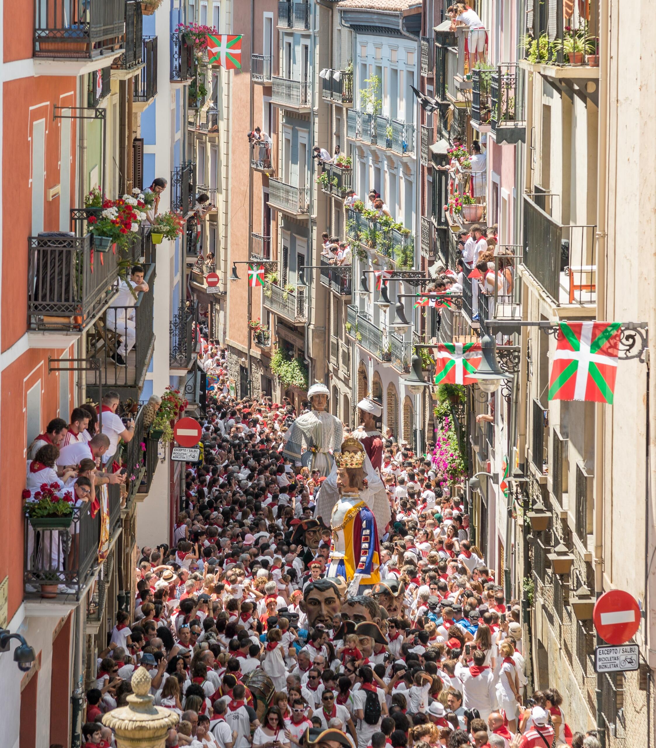 Una calle bulliciosa en pamplona en san fermin con un ambiente festivo, llena de multitudes con trajes blancos y vibrantes banderas vascas decorando los balcones.