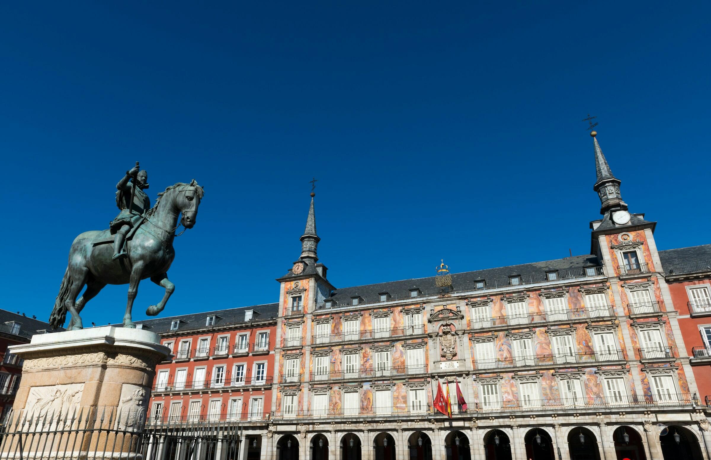 Una estatua ecuestre de bronce se alza en la plaza mayor de madrid frente a un edificio histórico con fachadas de colores, bajo un cielo azul despejado.