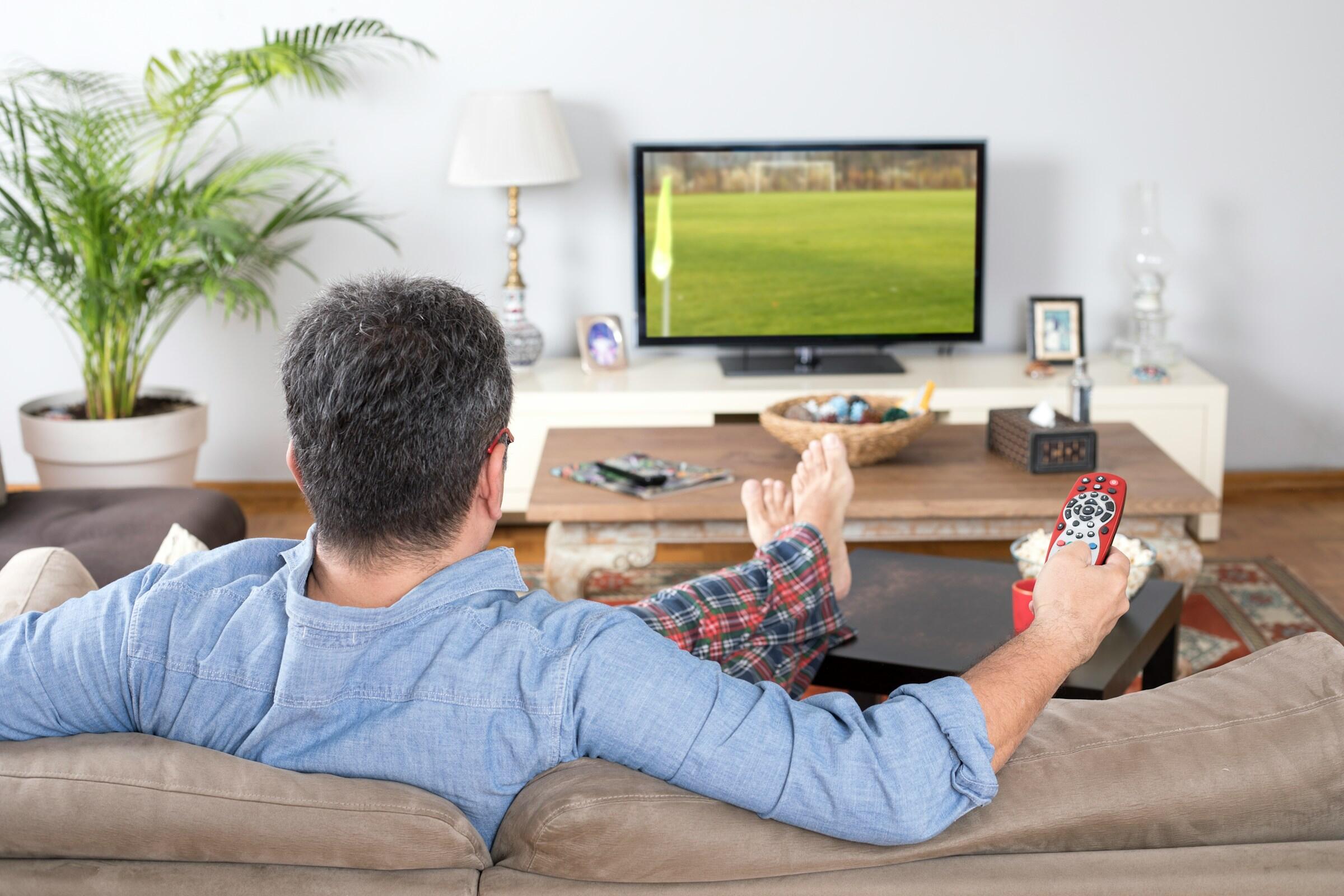 Un hombre vestido con ropa informal se relaja en un sofá viendo un partido de fútbol en la televisión mientras sujeta un mando a distancia.