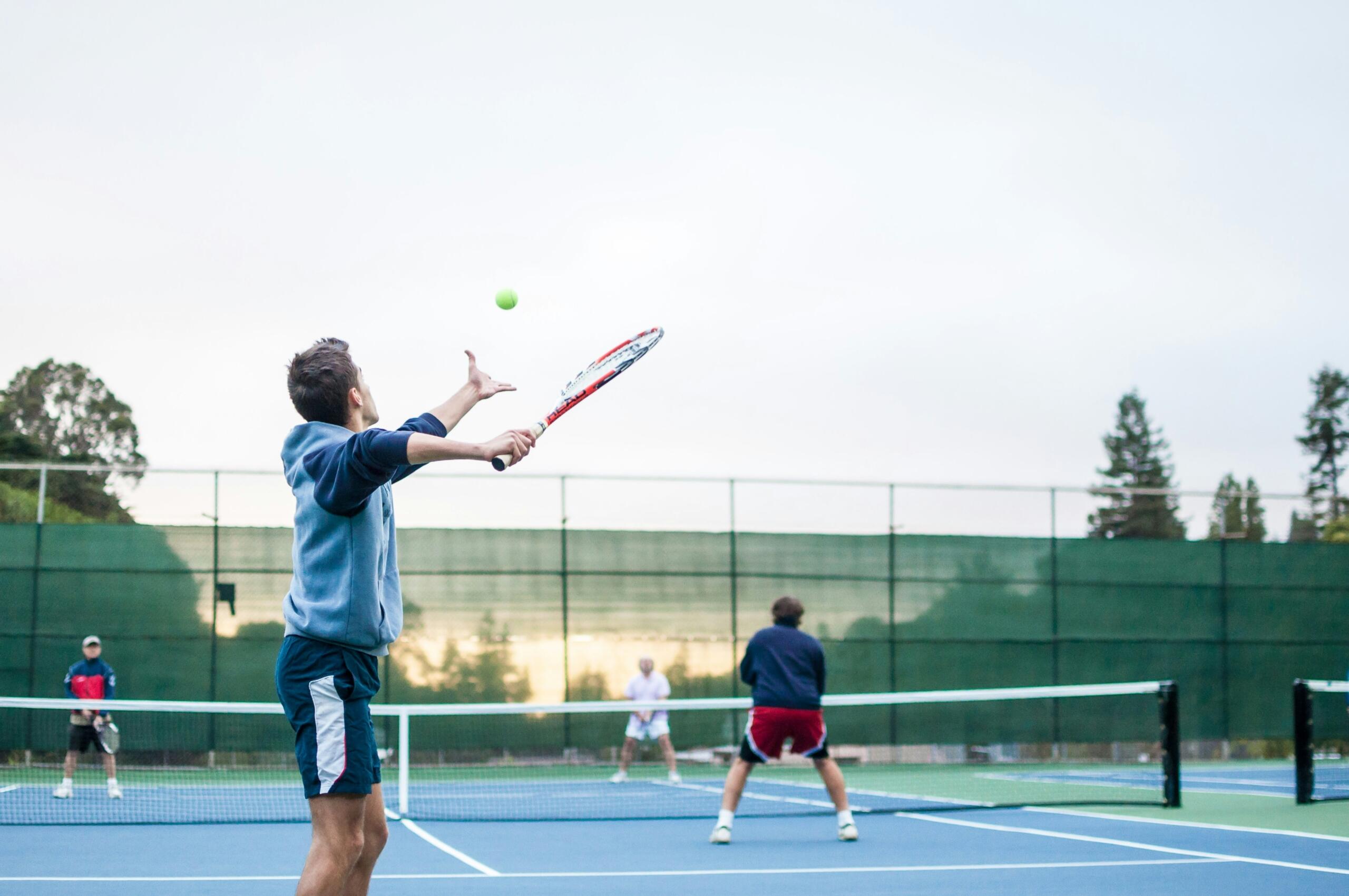 Un jugador de tenis saca una pelota en una pista al aire libre, con otros tres jugadores al fondo preparándose para el partido.