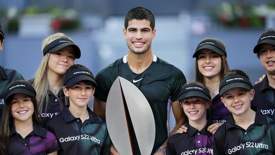 Un deportista sonriente sostiene un gran trofeo plateado, rodeado de jóvenes aficionados que llevan uniformes y gorras del Galaxy S22 Ultra, en un ambiente festivo y alegre.