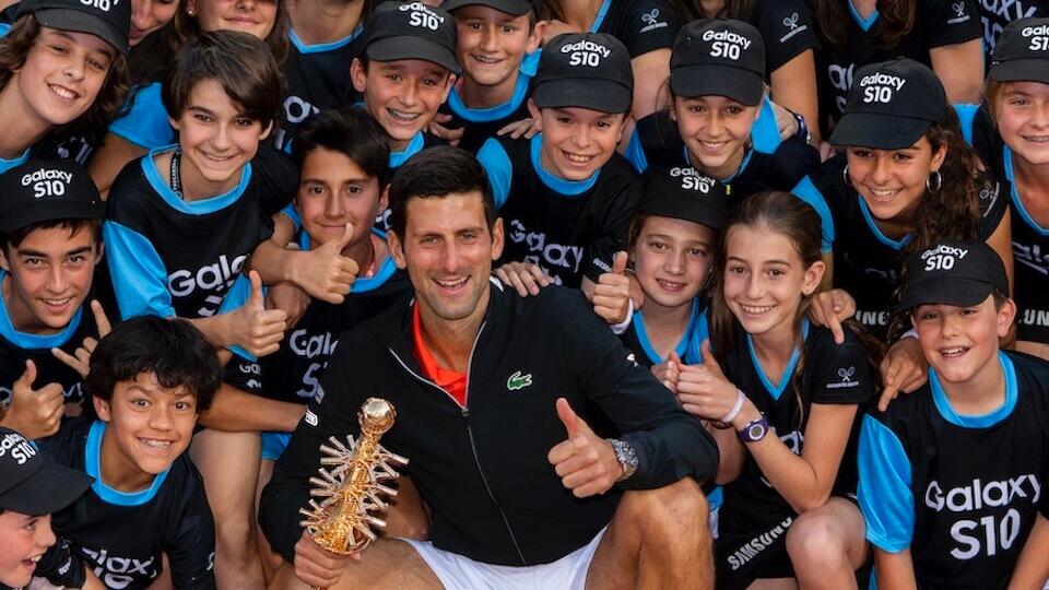 Un tenista sostiene un trofeo de oro, rodeado de niños sonrientes que llevan camisetas y gorras a juego del Galaxy S10, irradiando alegría y emoción.