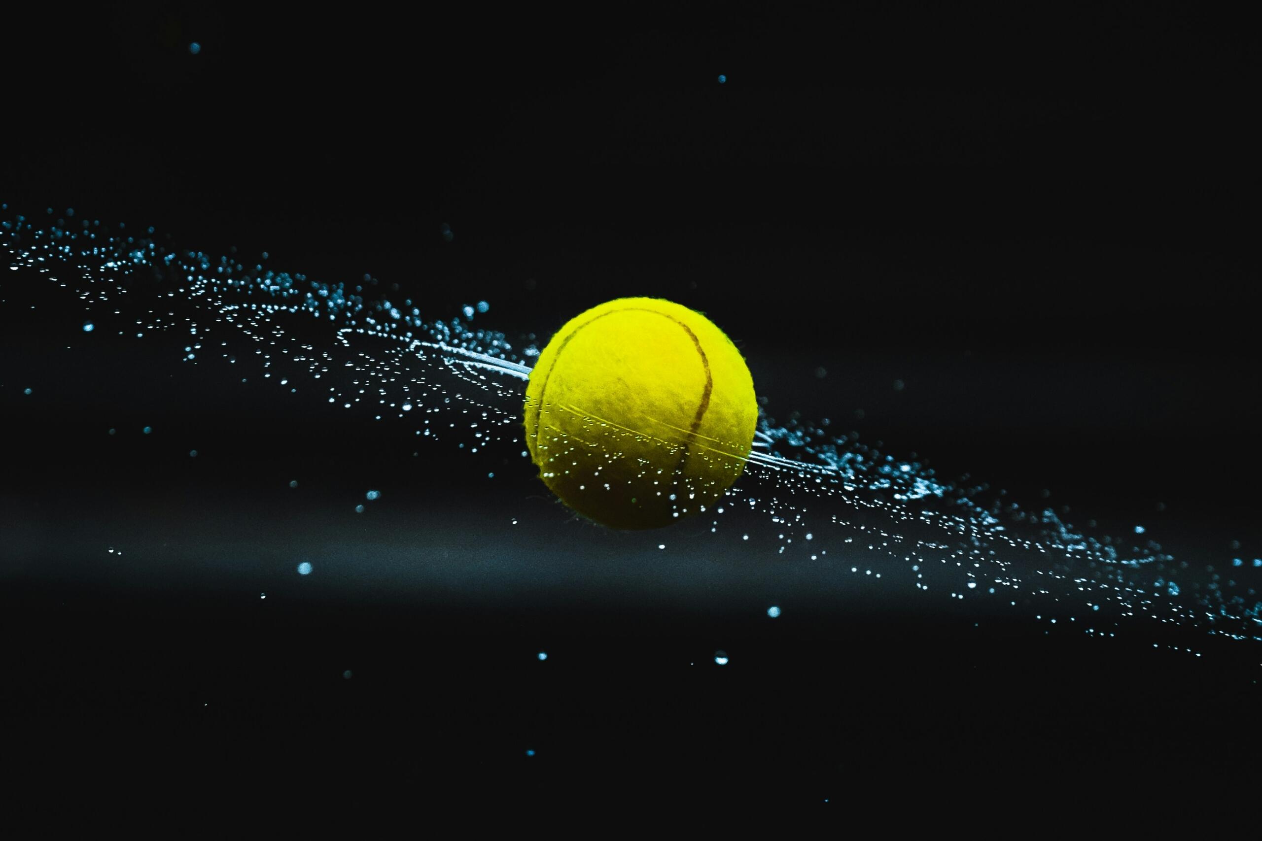 Una pelota de tenis amarilla brillante flotando en el aire, rodeada de un remolino de gotas azules sobre un fondo oscuro.
