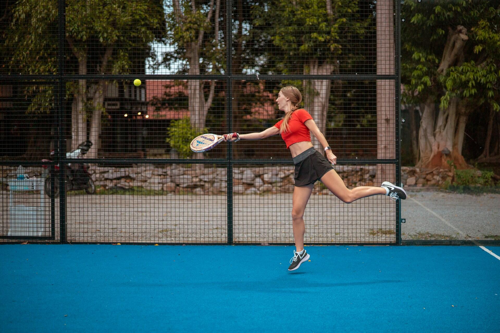 Mujer con camisa roja jugando pádel en una cancha al aire libre, sosteniendo una raqueta y enfocada en la pelota.