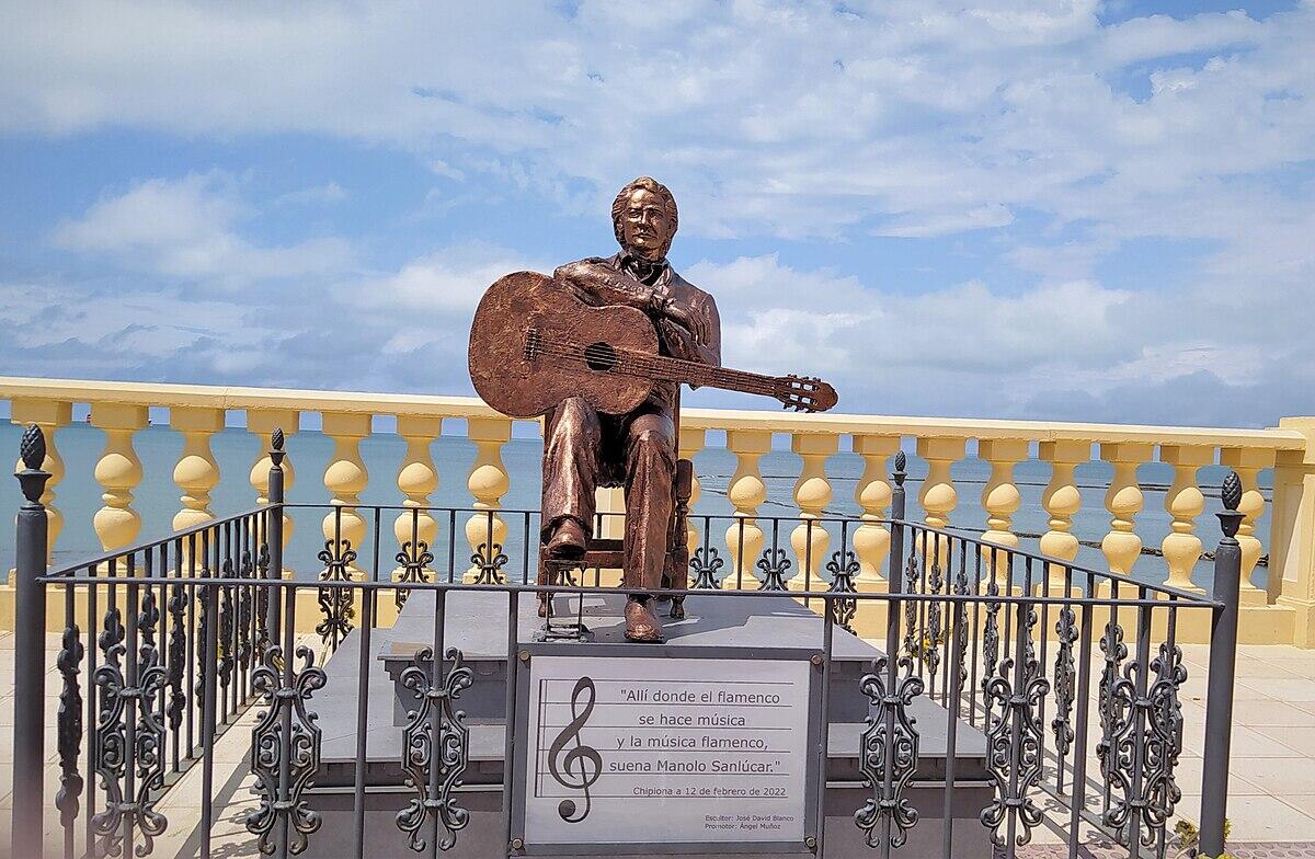 Estatua de bronce del guitarrista manolo sanlucar que aparece sentado, con vistas al mar, barandilla amarilla y cielo azul nublado de fondo en chipiona