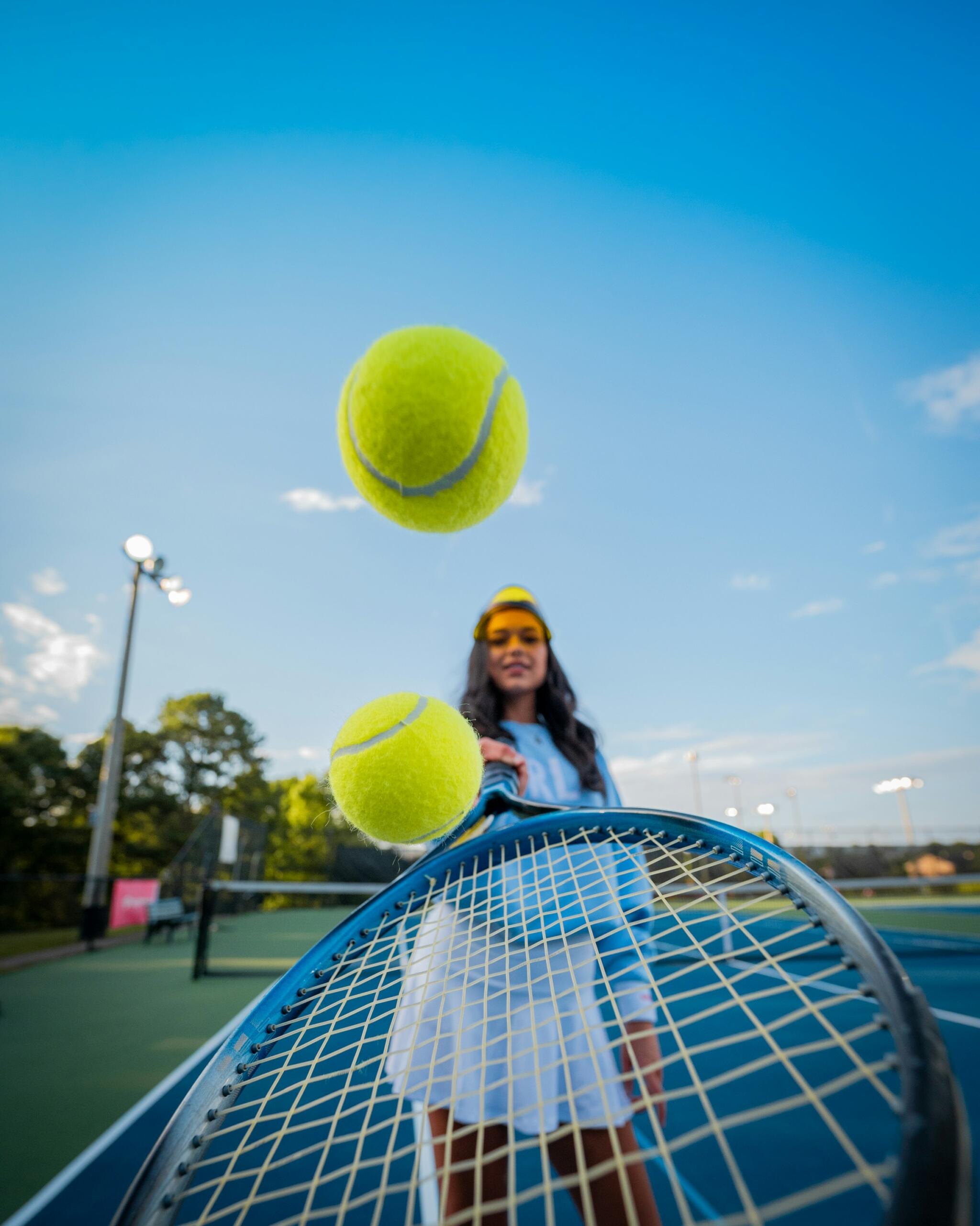 Una jugadors sostiene una raqueta de tenis con pelotas de tenis suspendidas en el aire contra un cielo azul despejado en una pista de tenis.