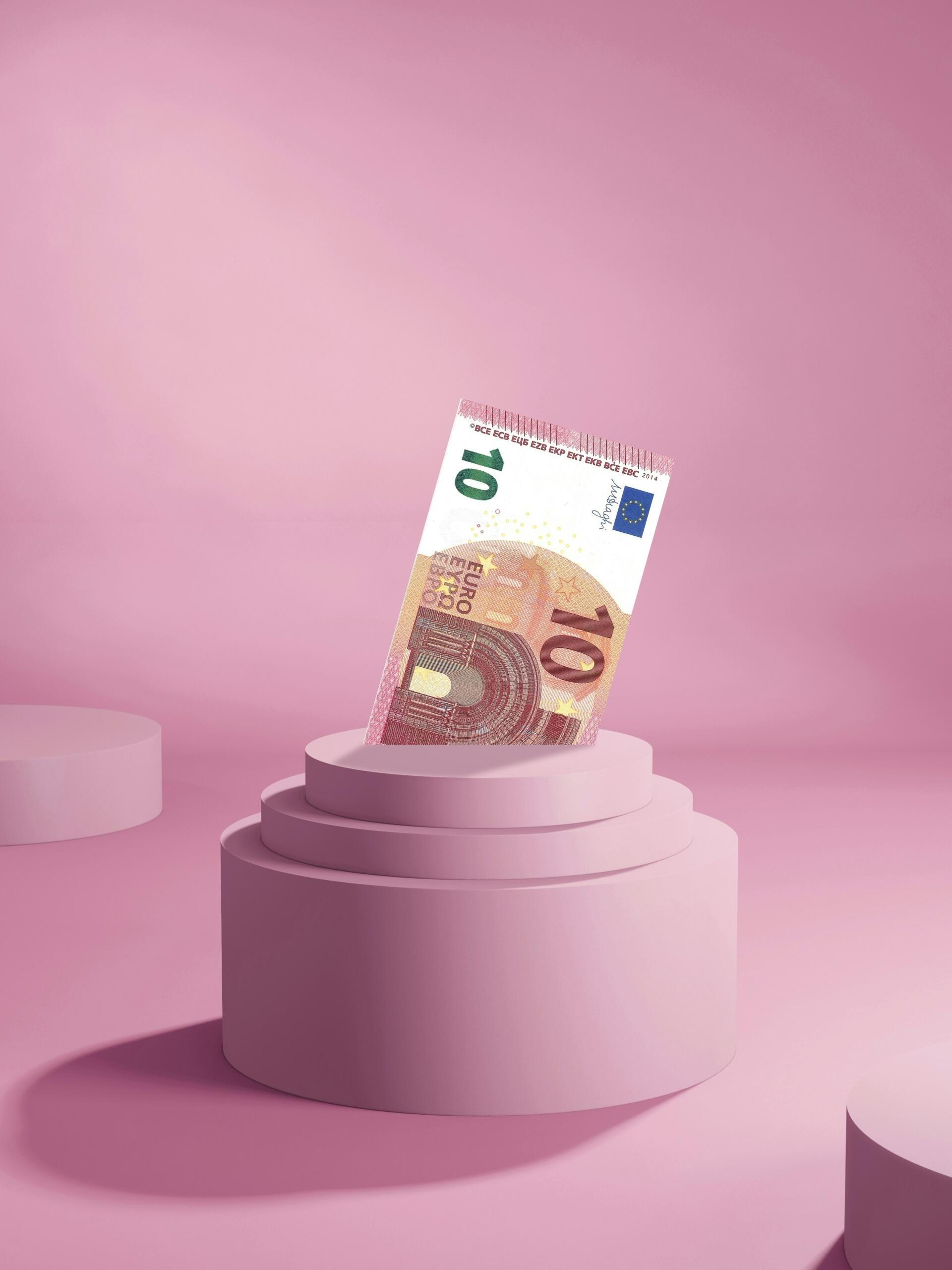 Un billete de 10 euros se alza sobre un pedestal circular rosa, sobre un fondo rosa suave.
