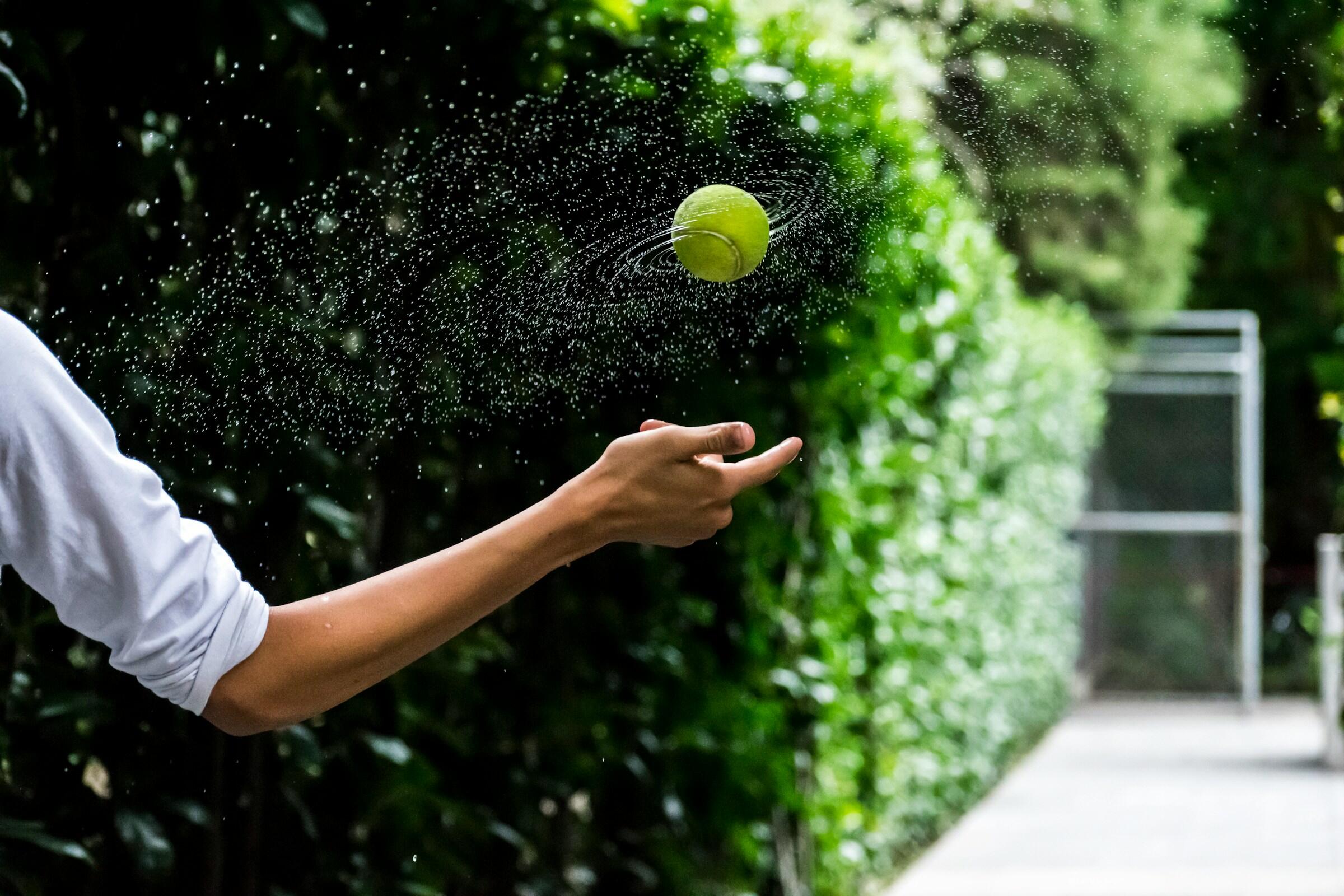Una mano lanza una pelota de tenis, creando una salpicadura de gotas de agua sobre un fondo verde vibrante de follaje.