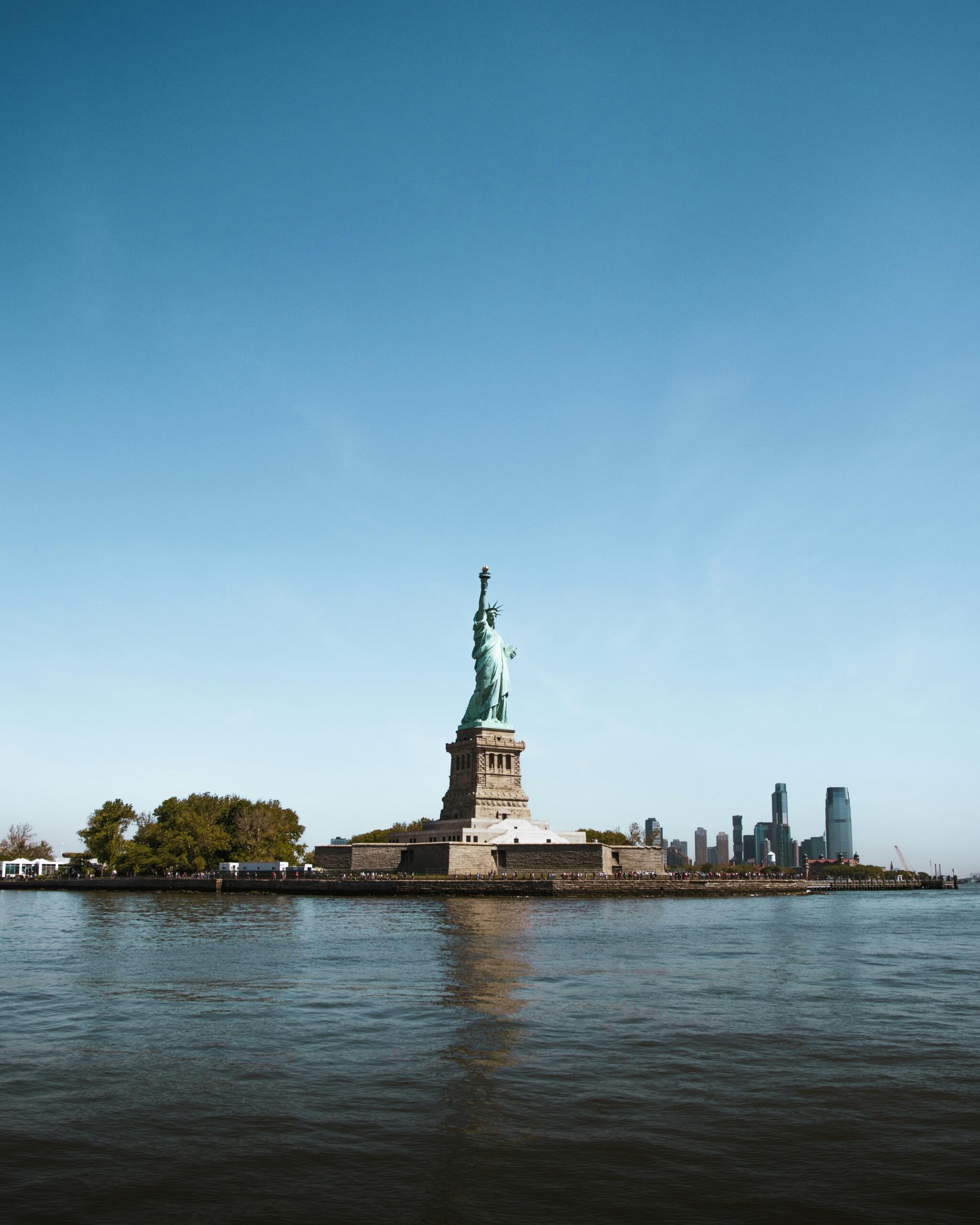 La Estatua de la Libertad se alza en la Isla de la Libertad, rodeada de agua, con el perfil de Nueva York al fondo.