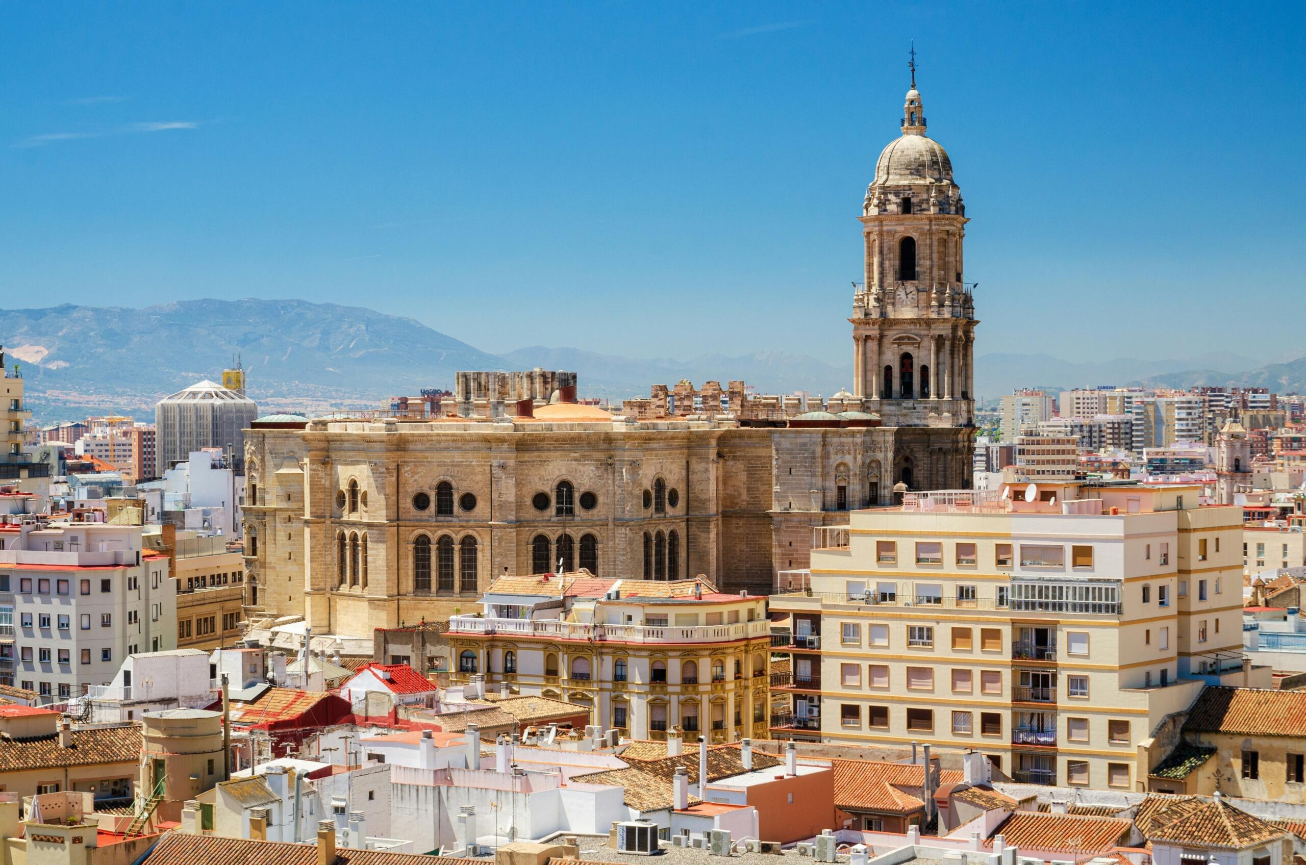 Una vista panorámica de Málaga con la Catedral y coloridos edificios en los tejados, con montañas al fondo bajo un cielo azul despejado.