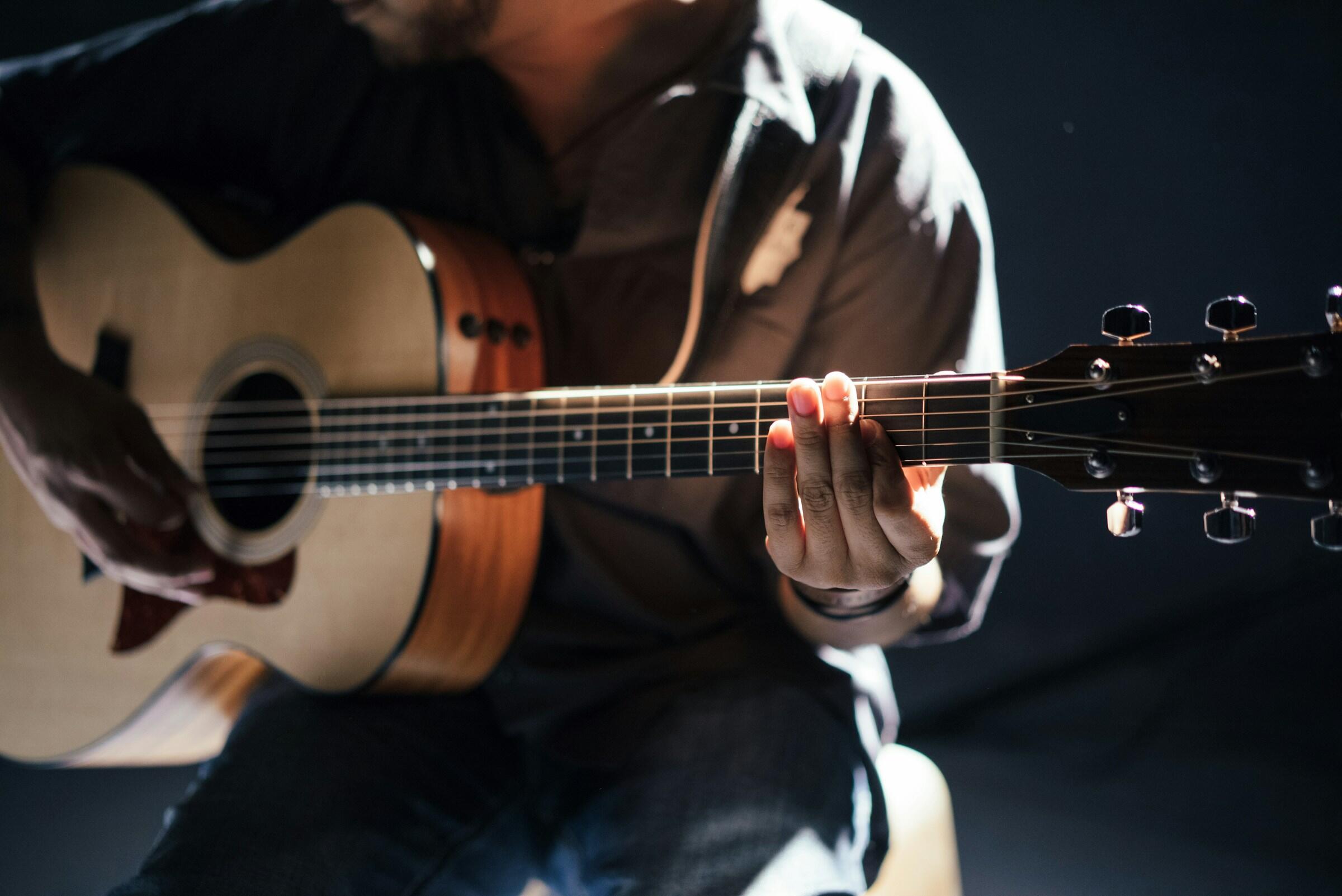 Primer plano de una persona tocando una guitarra acústica, destacando sus manos sobre el diapasón, en un entorno poco iluminado.