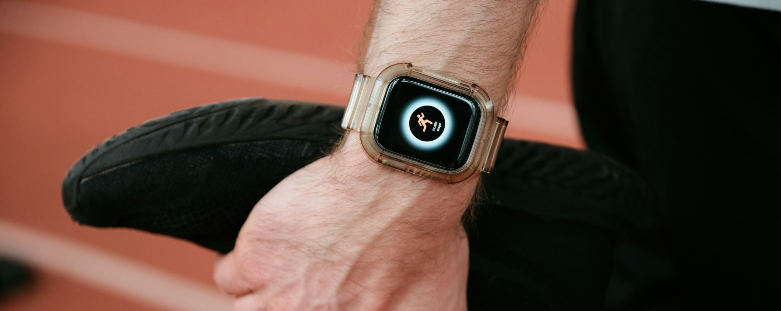 Primer plano de una mano con un reloj inteligente transparente que muestra un icono de carrera, junto a una zapatilla deportiva negra en una pista.