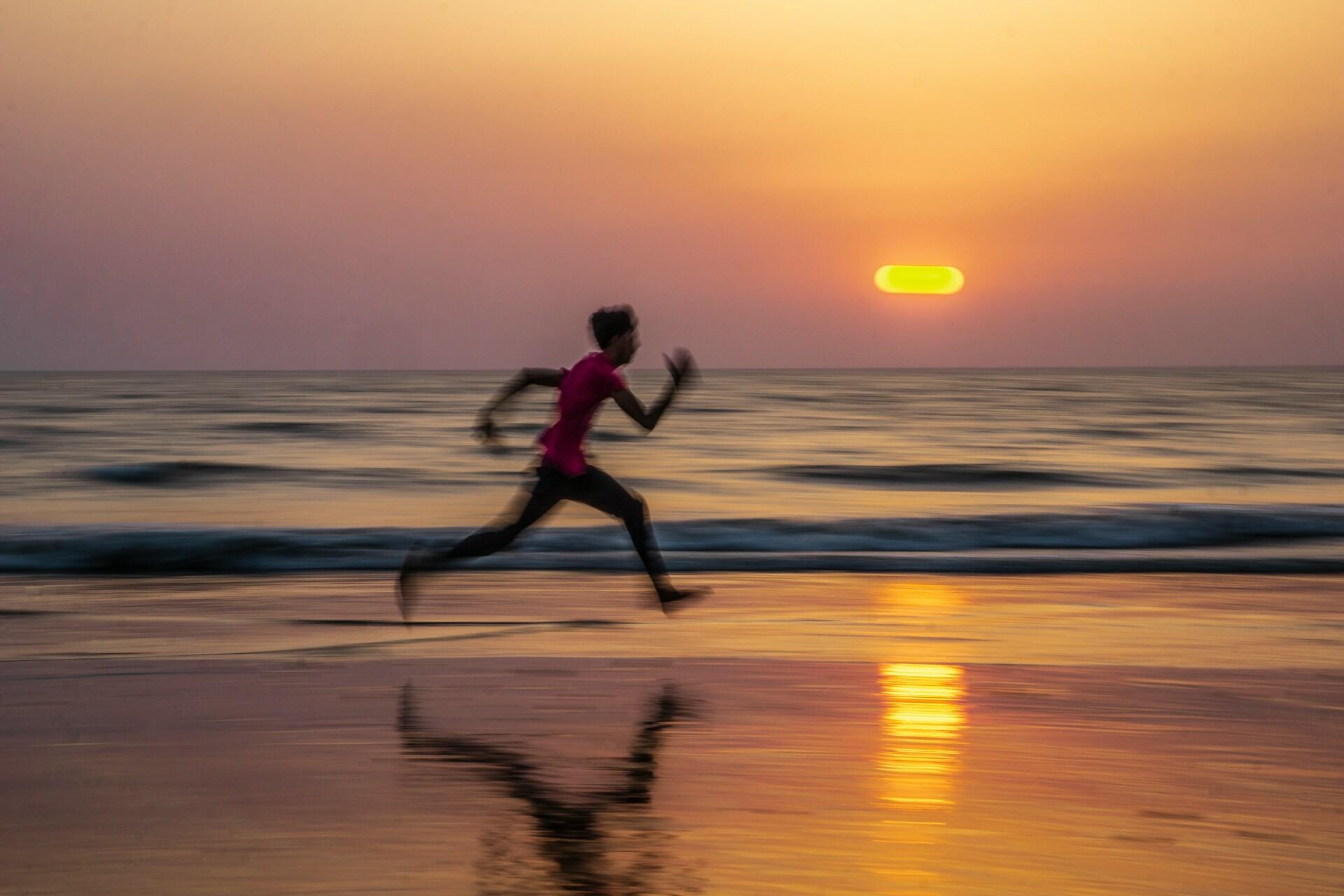 Un joven con una camiseta rosa corre por la playa al atardecer, con reflejos en la arena mojada y las olas de fondo.