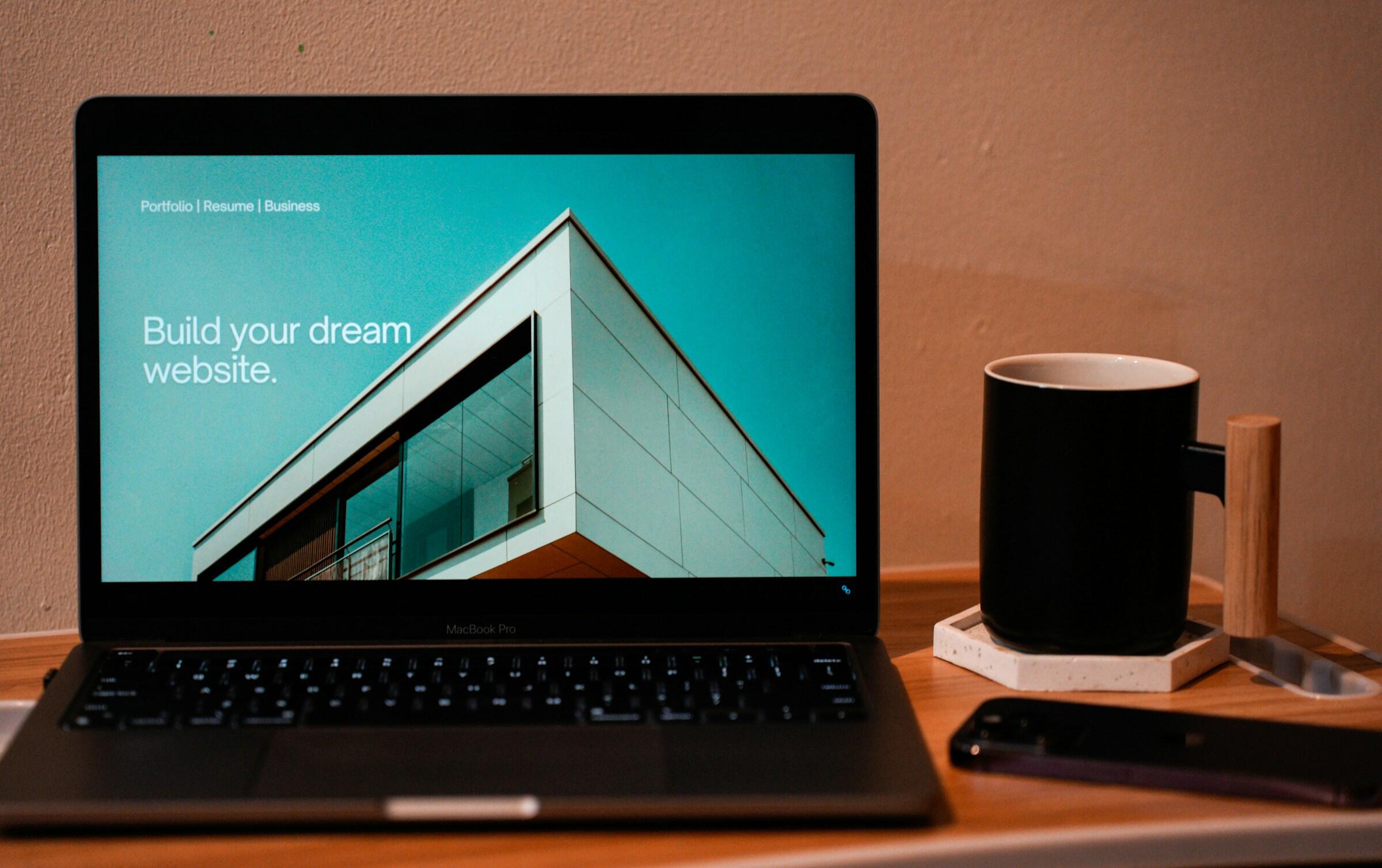 Un MacBook Pro que muestra una interfaz de diseño de sitios web con el texto «Construye el sitio web de tus sueños», junto a una taza negra y un smartphone.