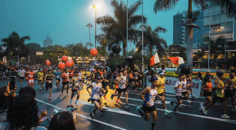 Una escena llena de energía de una maratón con corredores compitiendo, globos de colores y gente mirando el evento bajo un cielo nublado.