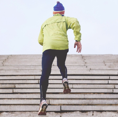 Una persona con una chaqueta amarilla brillante y un sombrero azul sube corriendo unos escalones de piedra, resaltando su atletismo y movimiento contra un cielo nublado.