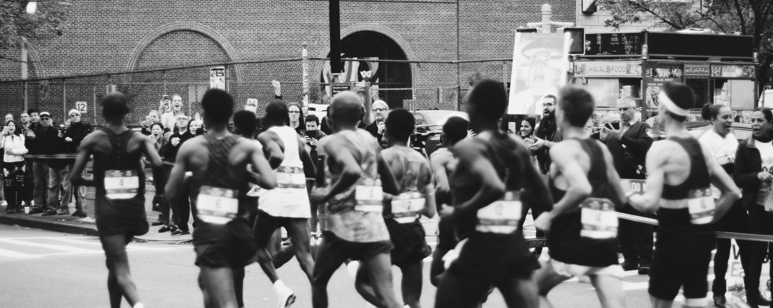 Un grupo de corredores de maratón, con dorsales, pasan corriendo entre espectadores que los animan en un entorno urbano en blanco y negro.