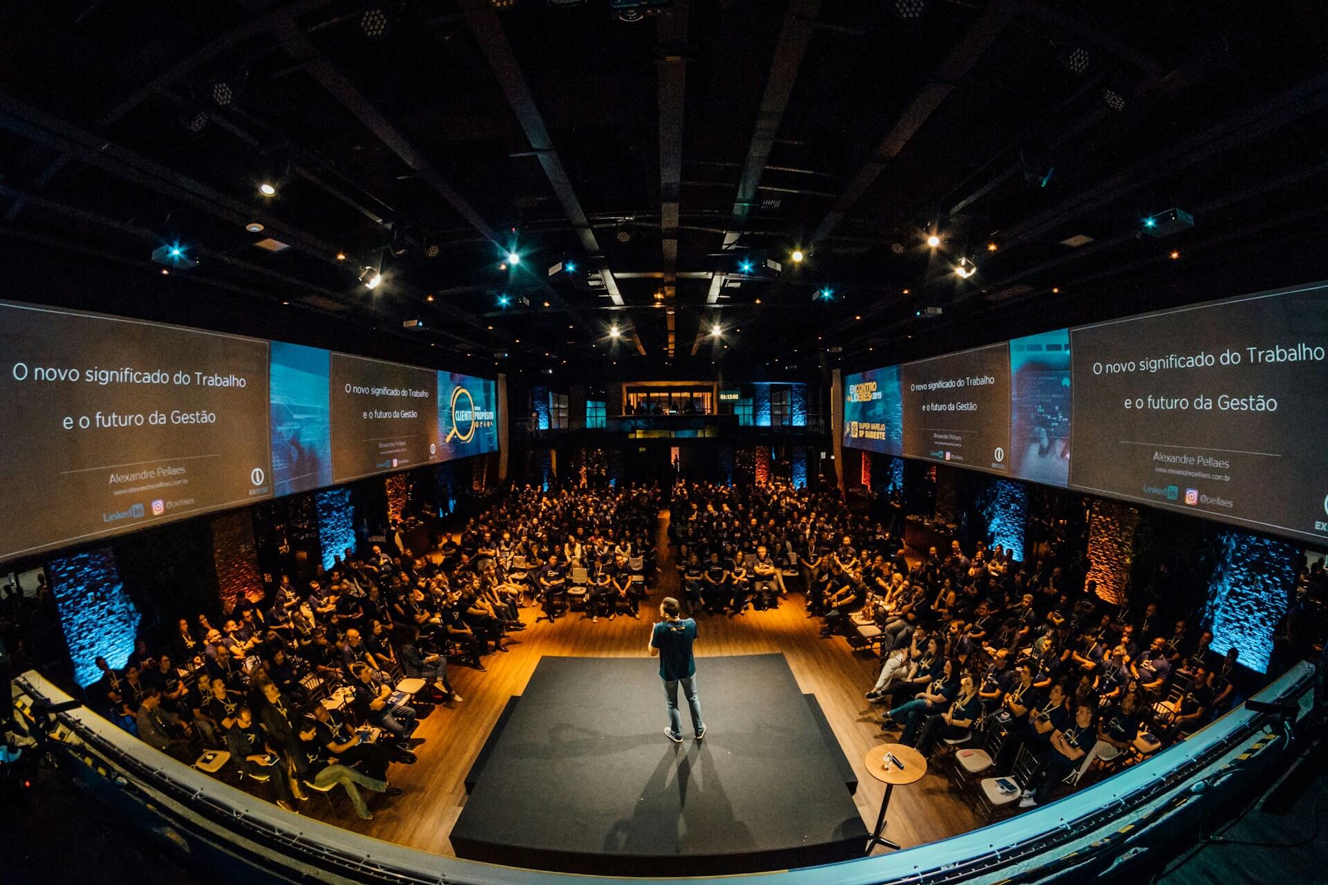 Un orador se dirige a una gran audiencia en un evento sobre el futuro del trabajo y la gestión, con presentaciones mostradas en pantallas.