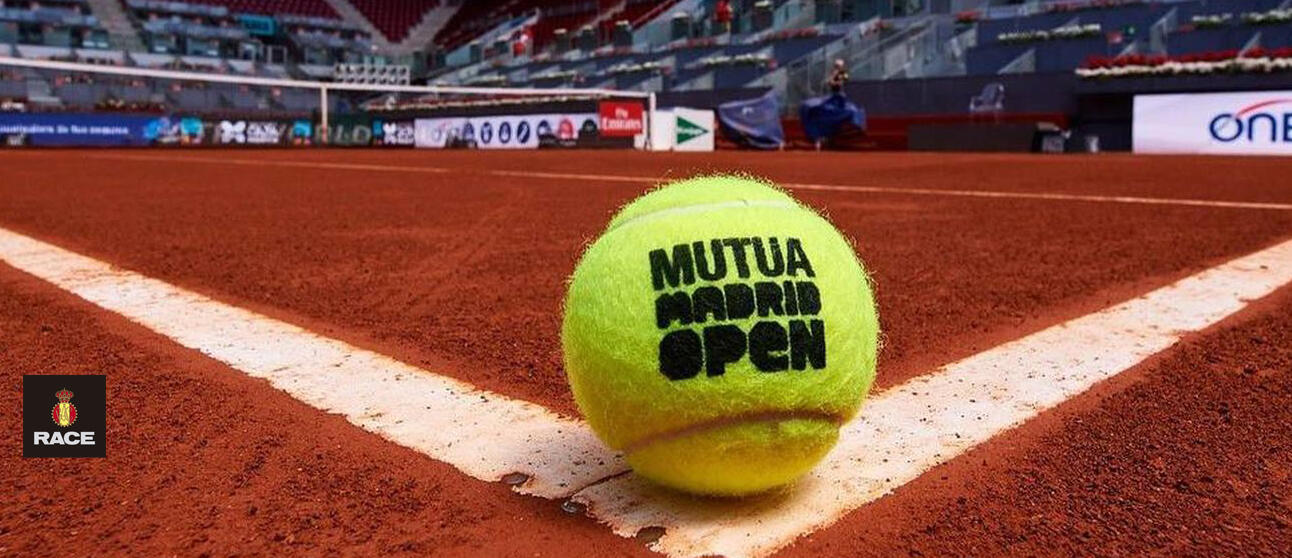 Primer plano de una pelota de tenis amarilla con la inscripción «Mutua Madrid Open» sobre una pista de tierra batida roja. Las líneas blancas enmarcan la escena, con las gradas y las pancartas difuminadas al fondo.