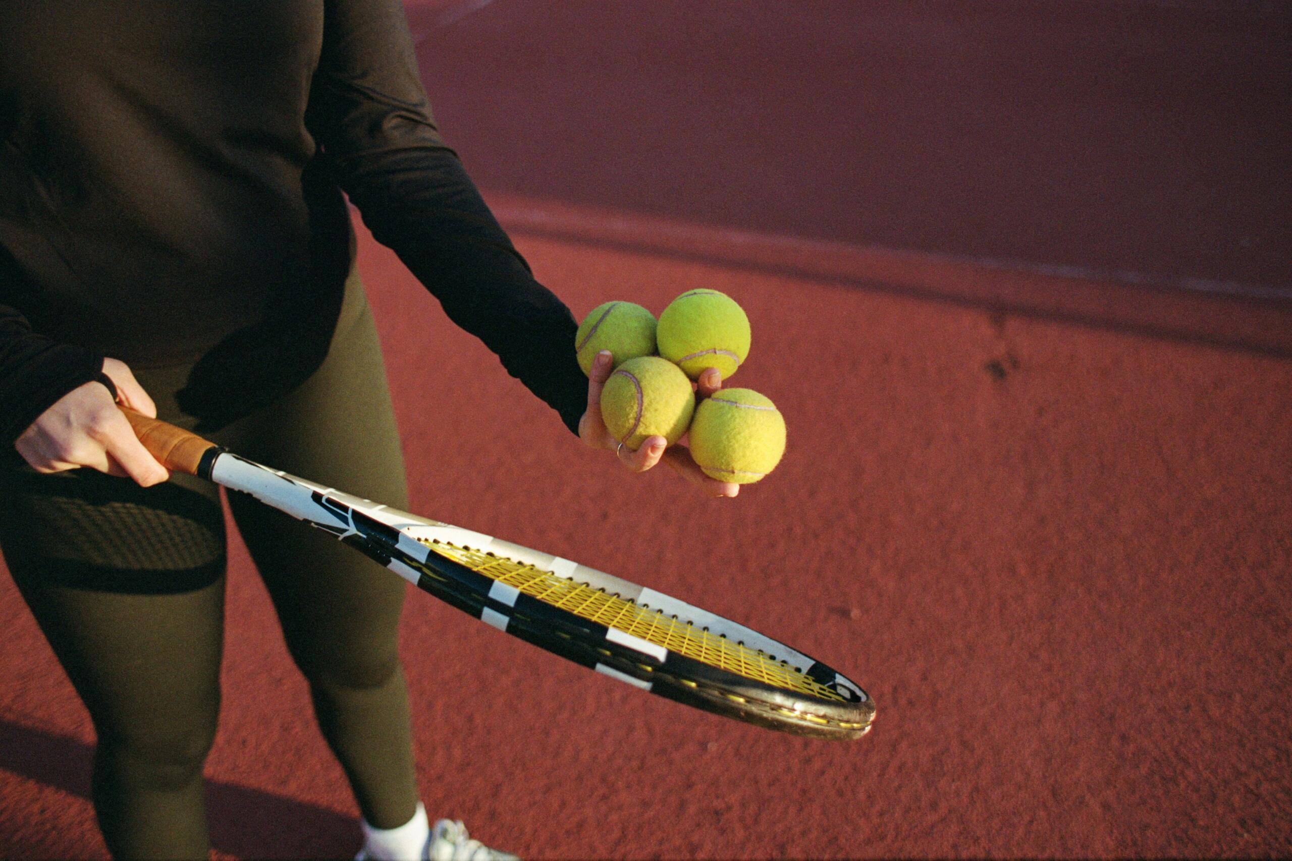 Un jugador con mallas negras sostiene una raqueta de tenis en una mano y cuatro pelotas de tenis verdes en la otra en una pista roja.