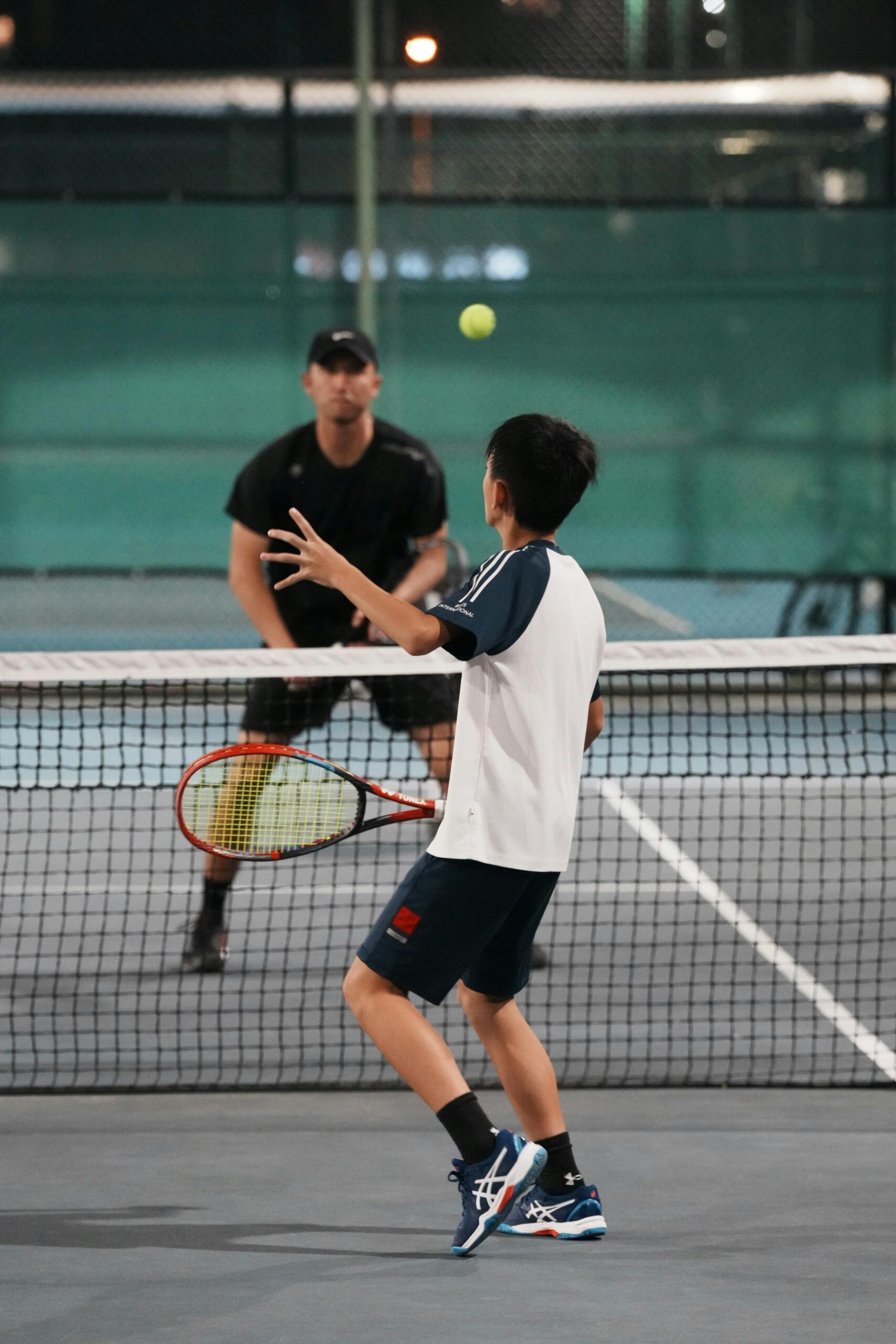 Un chico joven juega al tenis, preparado para golpear una pelota mientras está de pie en una pista de noche, con una red entre él y otro jugador.