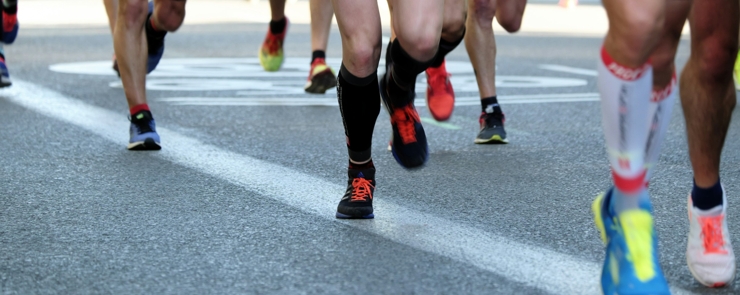 Primer plano de las piernas de varios corredores en movimiento durante una carrera, mostrando diferentes zapatillas deportivas y el efecto de movimiento borroso en una carretera asfaltada.