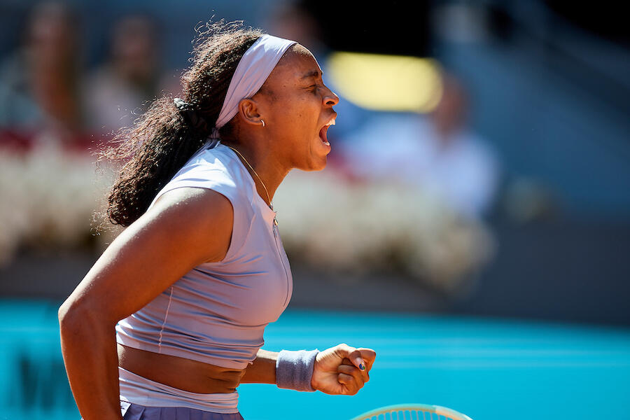 Un tenista en plena acción, con una cinta en la cabeza, celebra con el puño cerrado y una expresión de alegría, con una multitud borrosa al fondo.