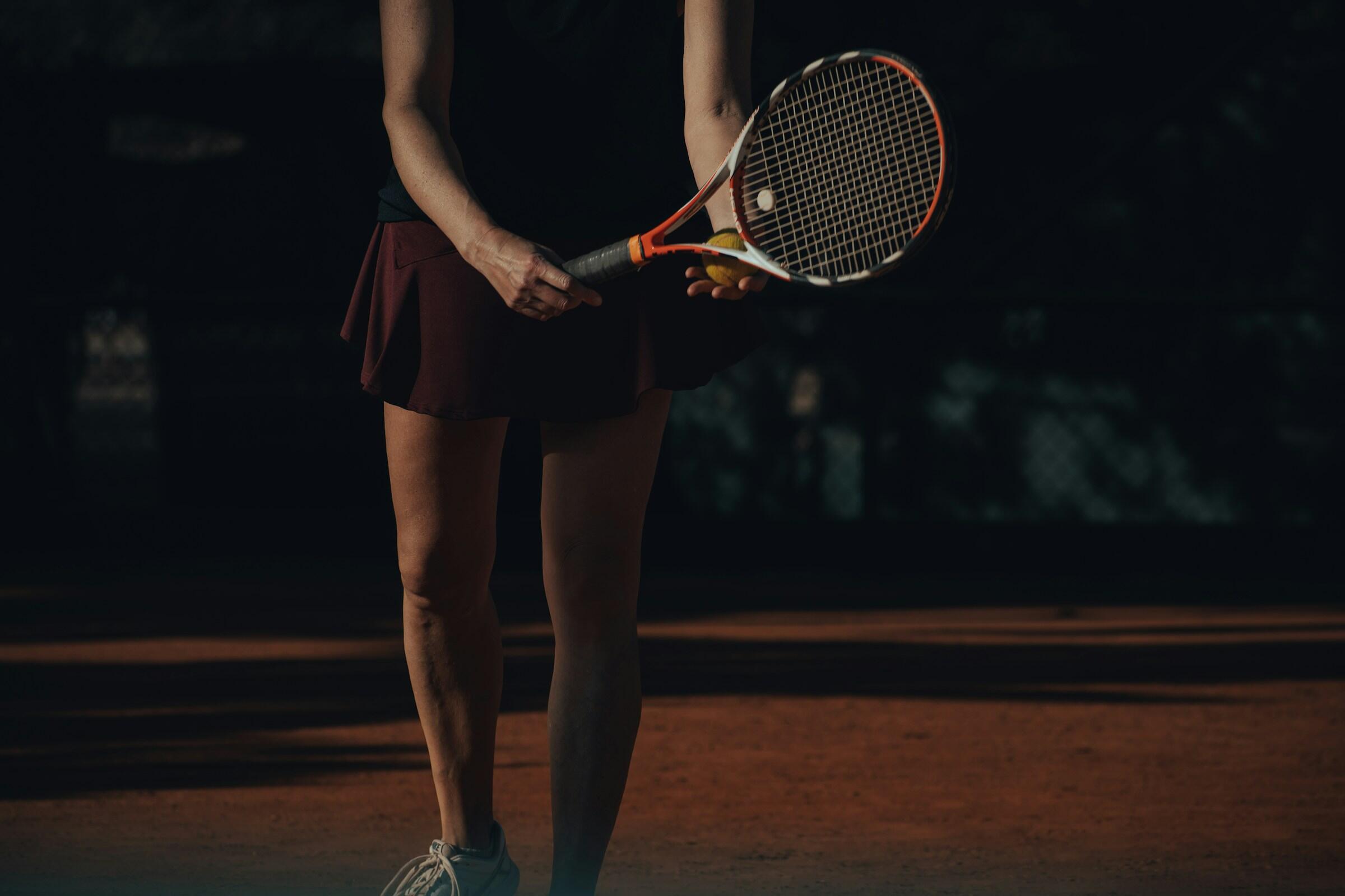 Una mujer con top negro y falda de tenis granate se prepara para servir en una pista de tierra batida, sujetando una raqueta y una pelota de tenis.