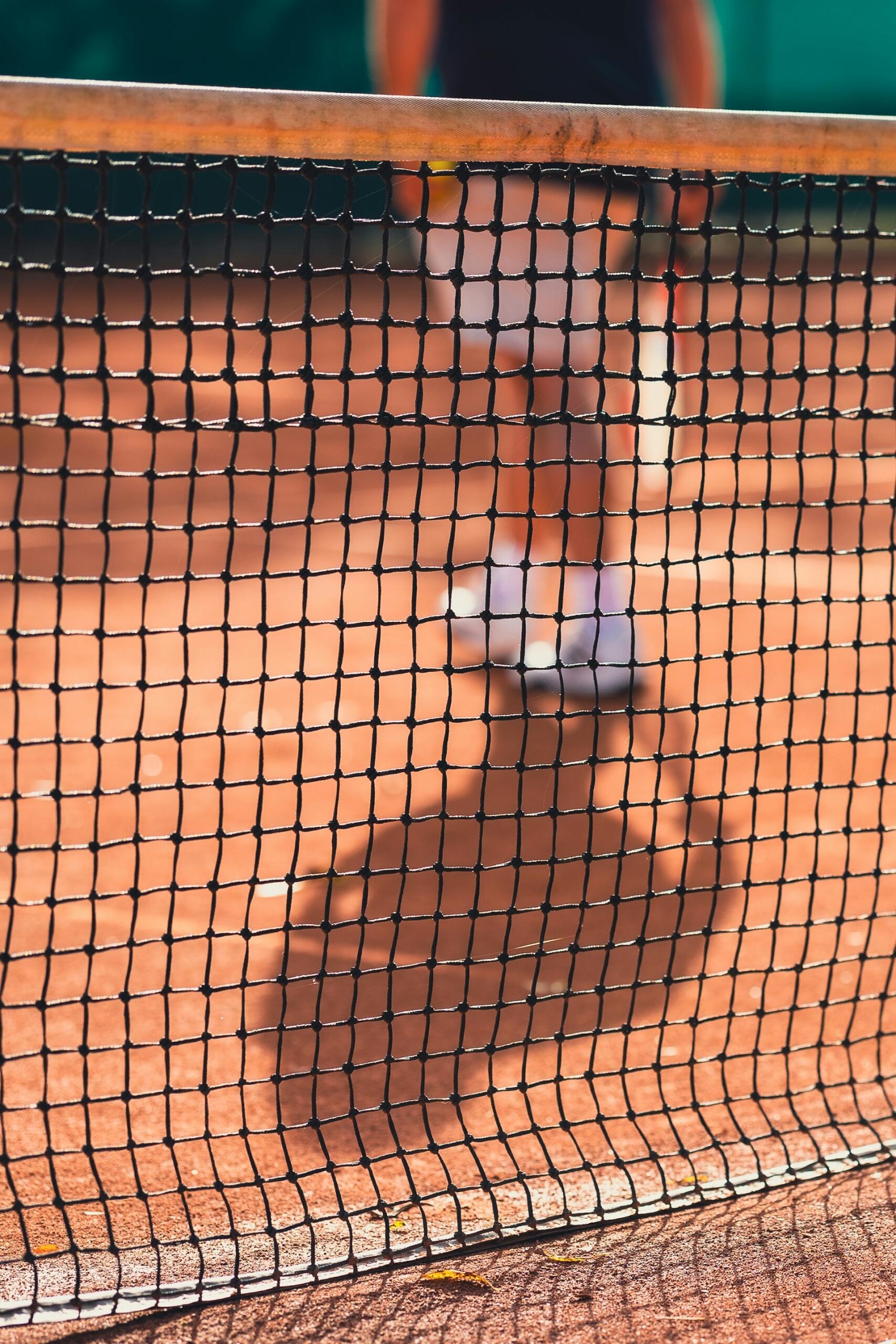 Escena en una pista de tenis con el foco puesto en la red; un jugador difuminado, vestido con pantalones cortos blancos y zapatillas deportivas, se encuentra de pie sobre la superficie de tierra batida, creando un ambiente relajado.