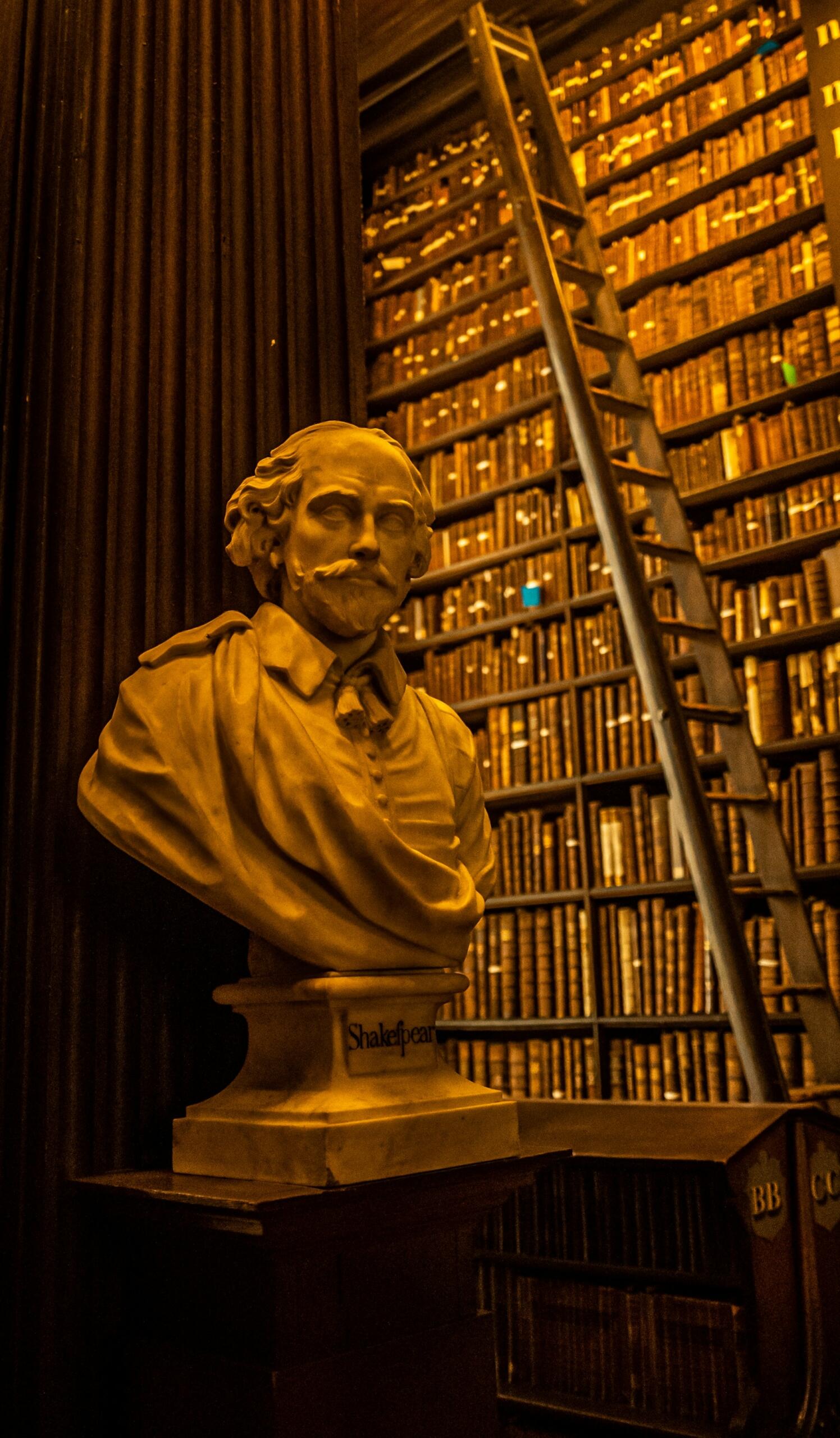 Un busto de mármol blanco de Shakespeare se alza prominentemente frente a una pared de estanterías antiguas, con una escalera de madera cerca.