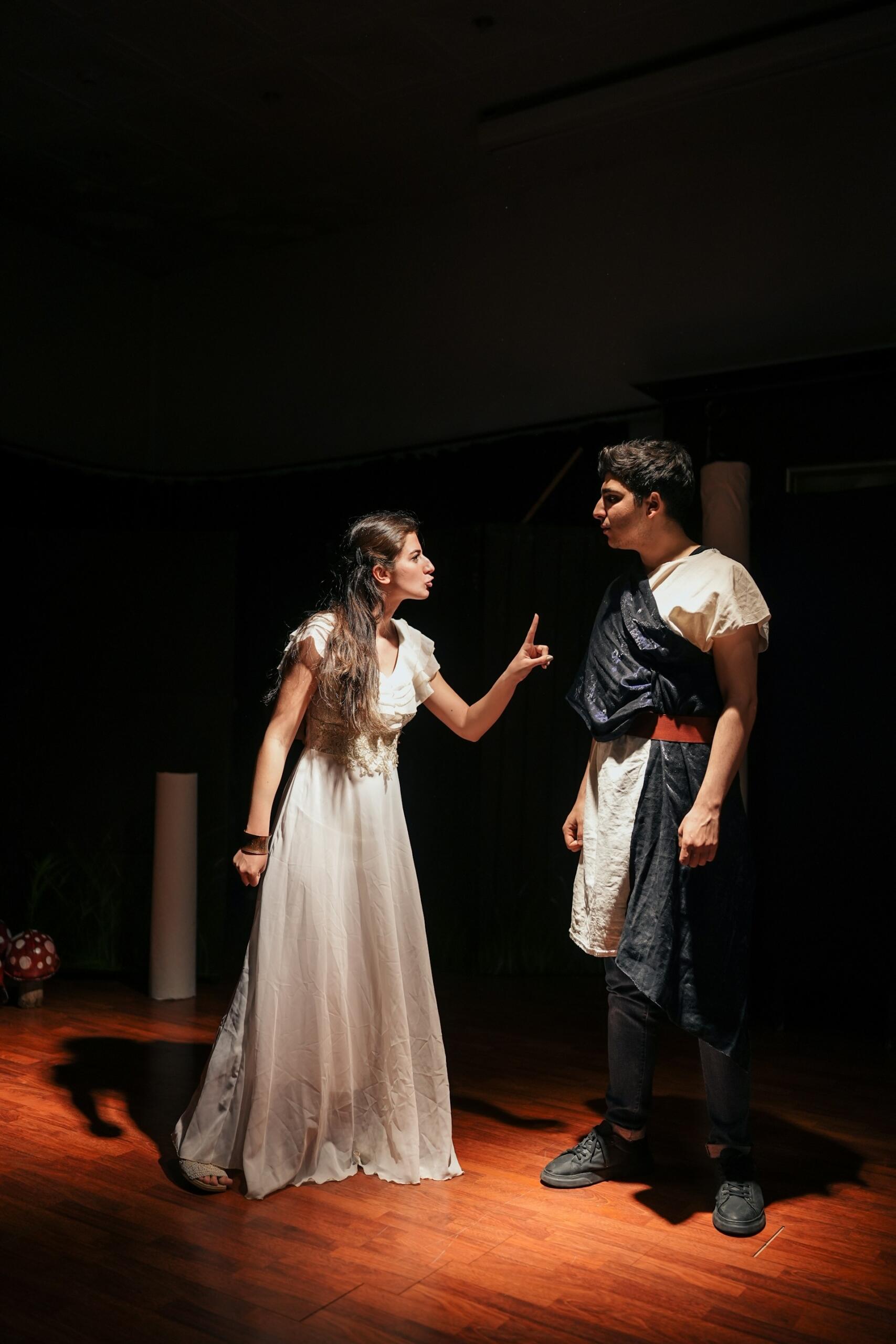 Una escena dramática en el escenario en la que aparece una mujer con un vestido blanco suelto que gesticula enfáticamente hacia un hombre vestido de oscuro.
