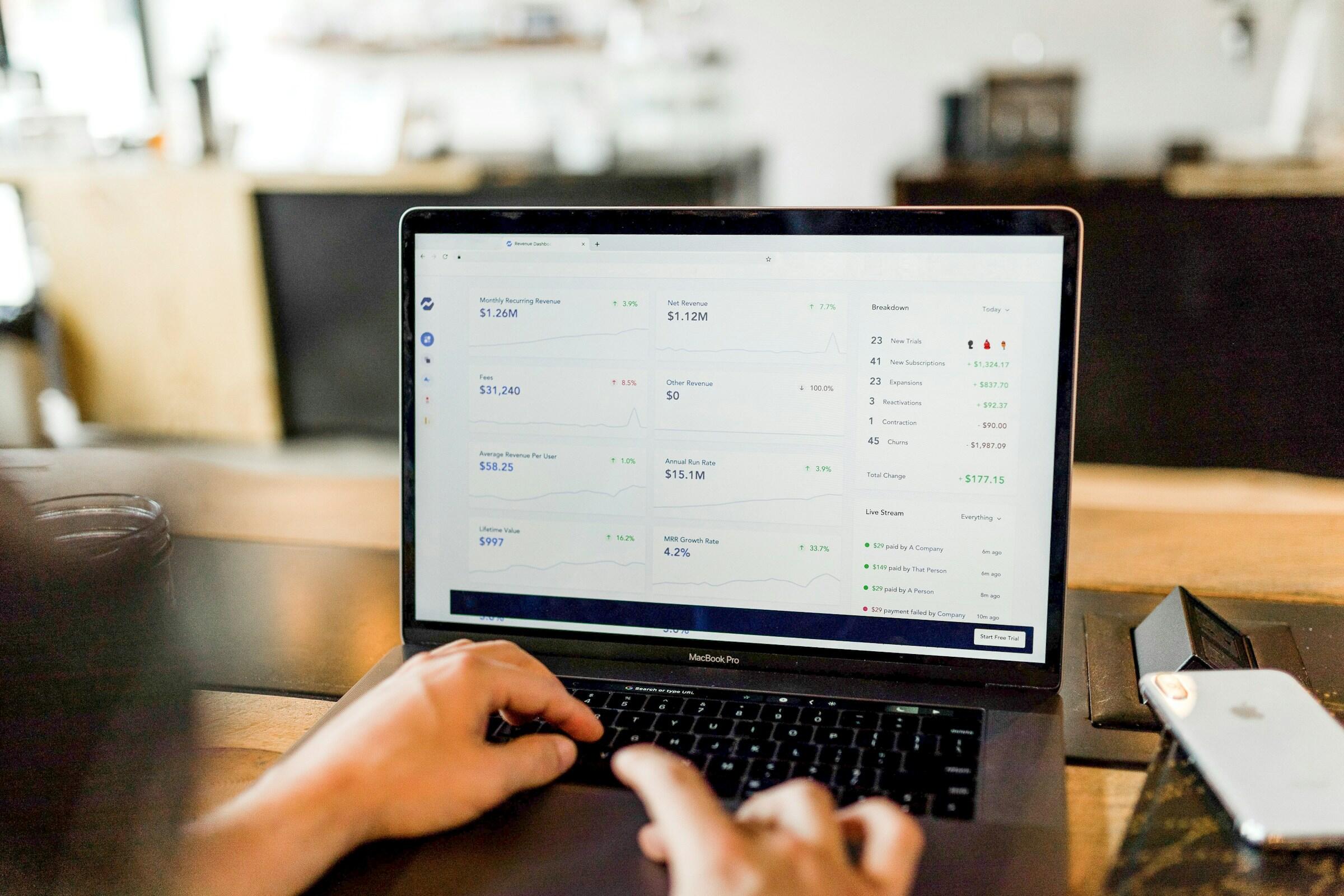 Una persona analiza los datos financieros en la pantalla de un portátil, mostrando métricas como los ingresos recurrentes mensuales y las estadísticas de usuarios en una cafetería.