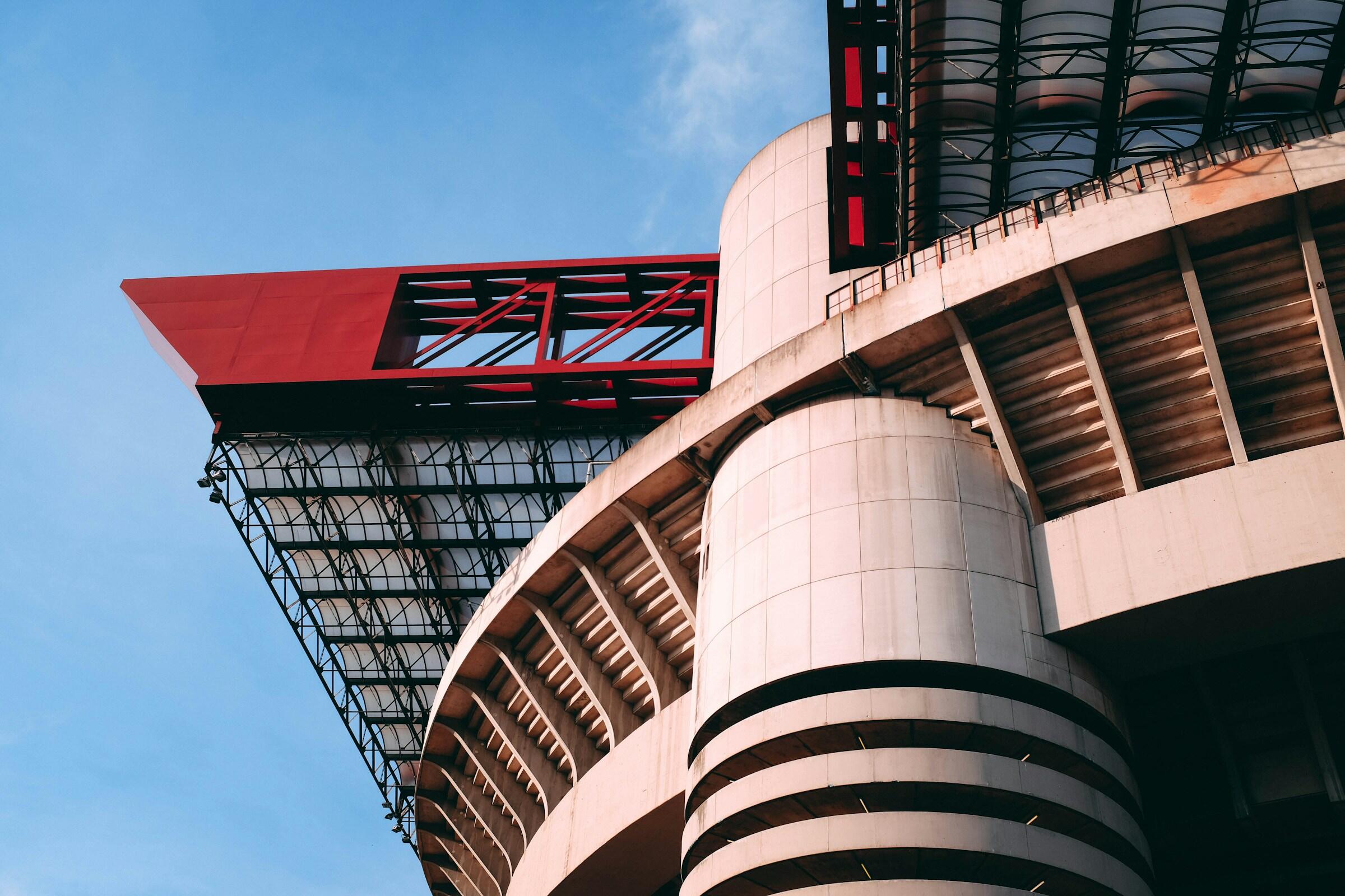 Primer plano de las características arquitectónicas de un estadio moderno, con columnas curvas de hormigón y un llamativo voladizo rojo sobre un cielo azul del estadio de san siro