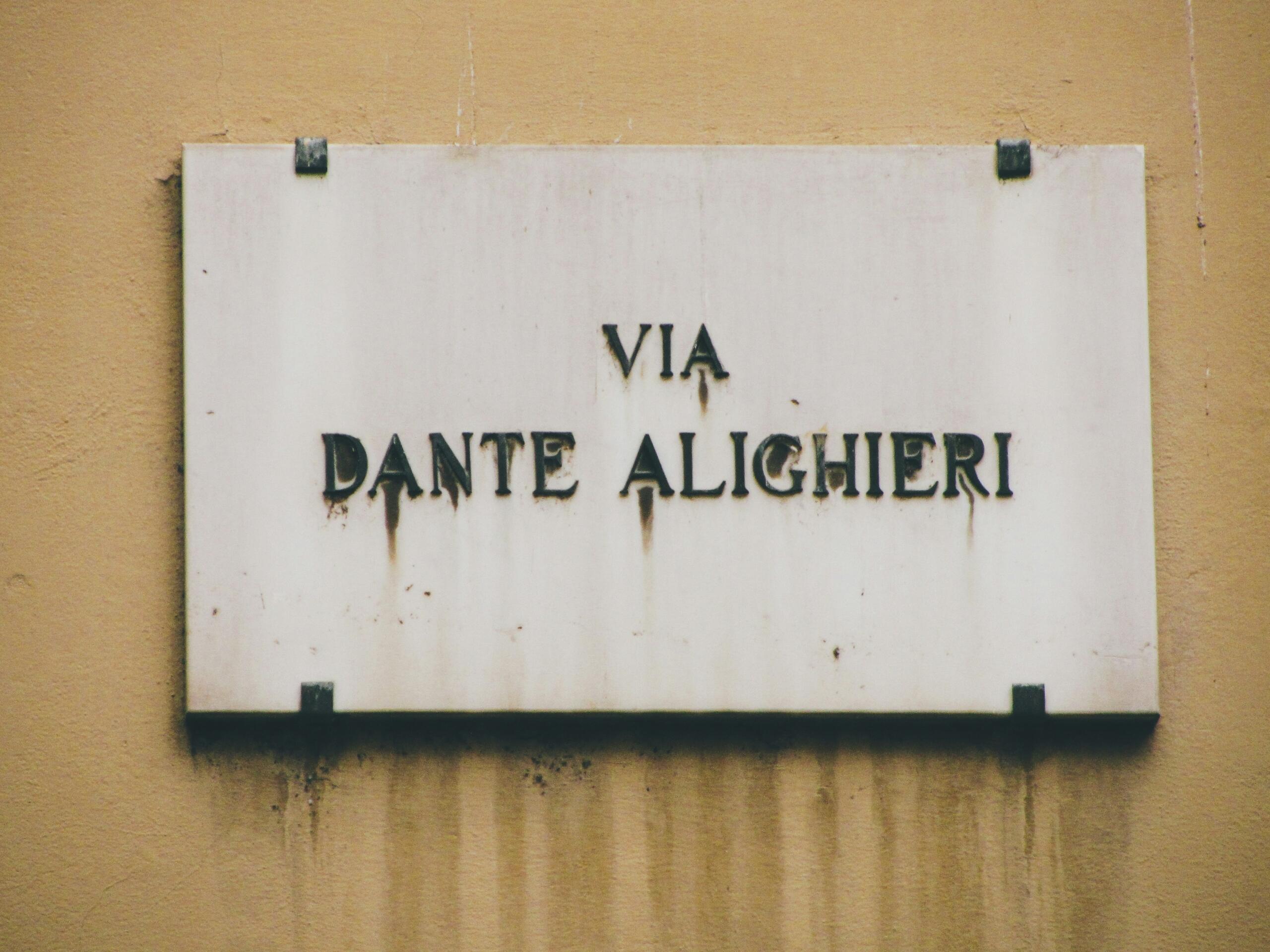 Placa de calle con la leyenda «Via Dante Alighieri», montada en una pared con un acabado rústico y desgastado.