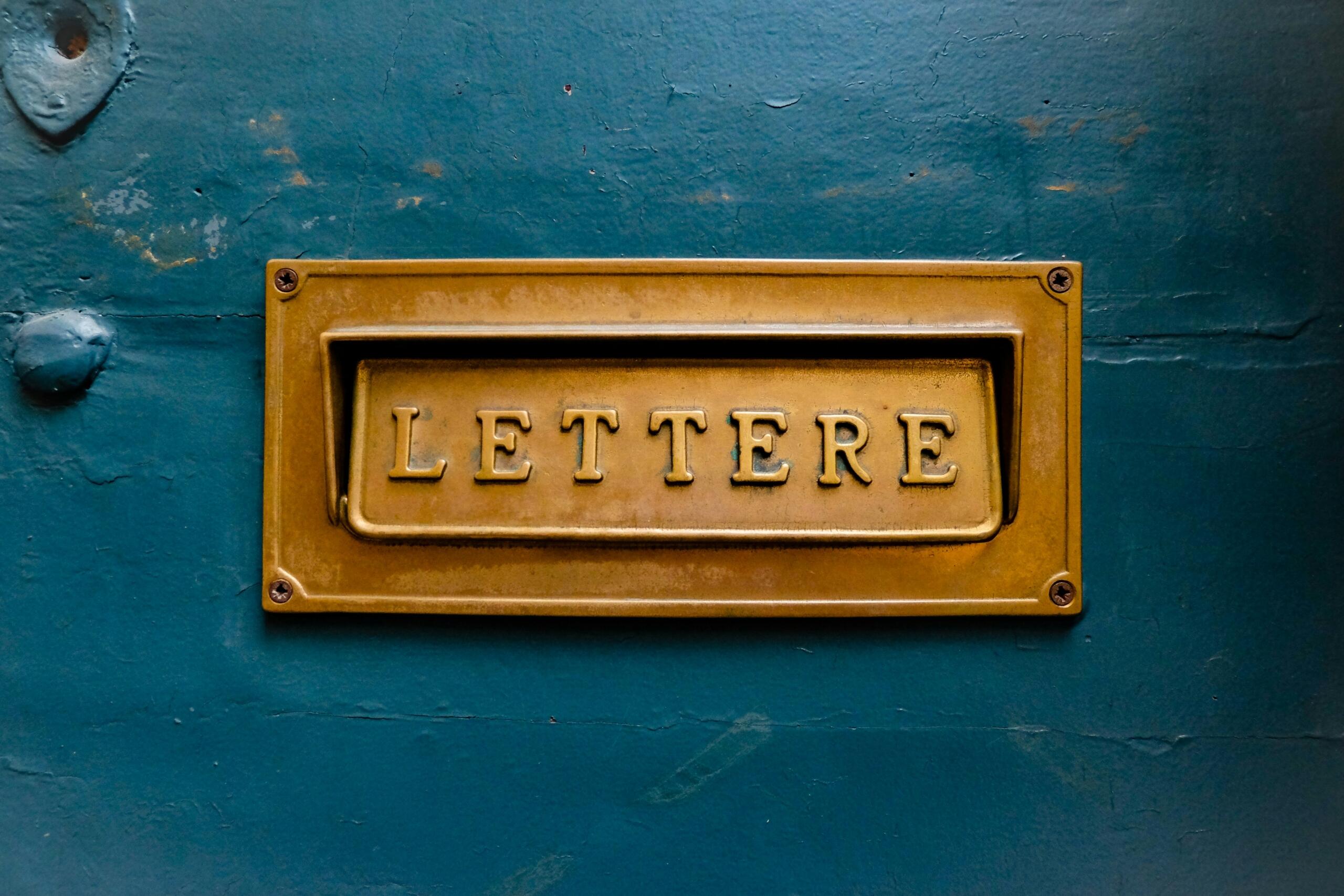 Primer plano de una placa de buzón de latón vintage con la etiqueta «LETTERE» sobre una pared de color verde azulado con textura, que muestra un diseño clásico y pátina.