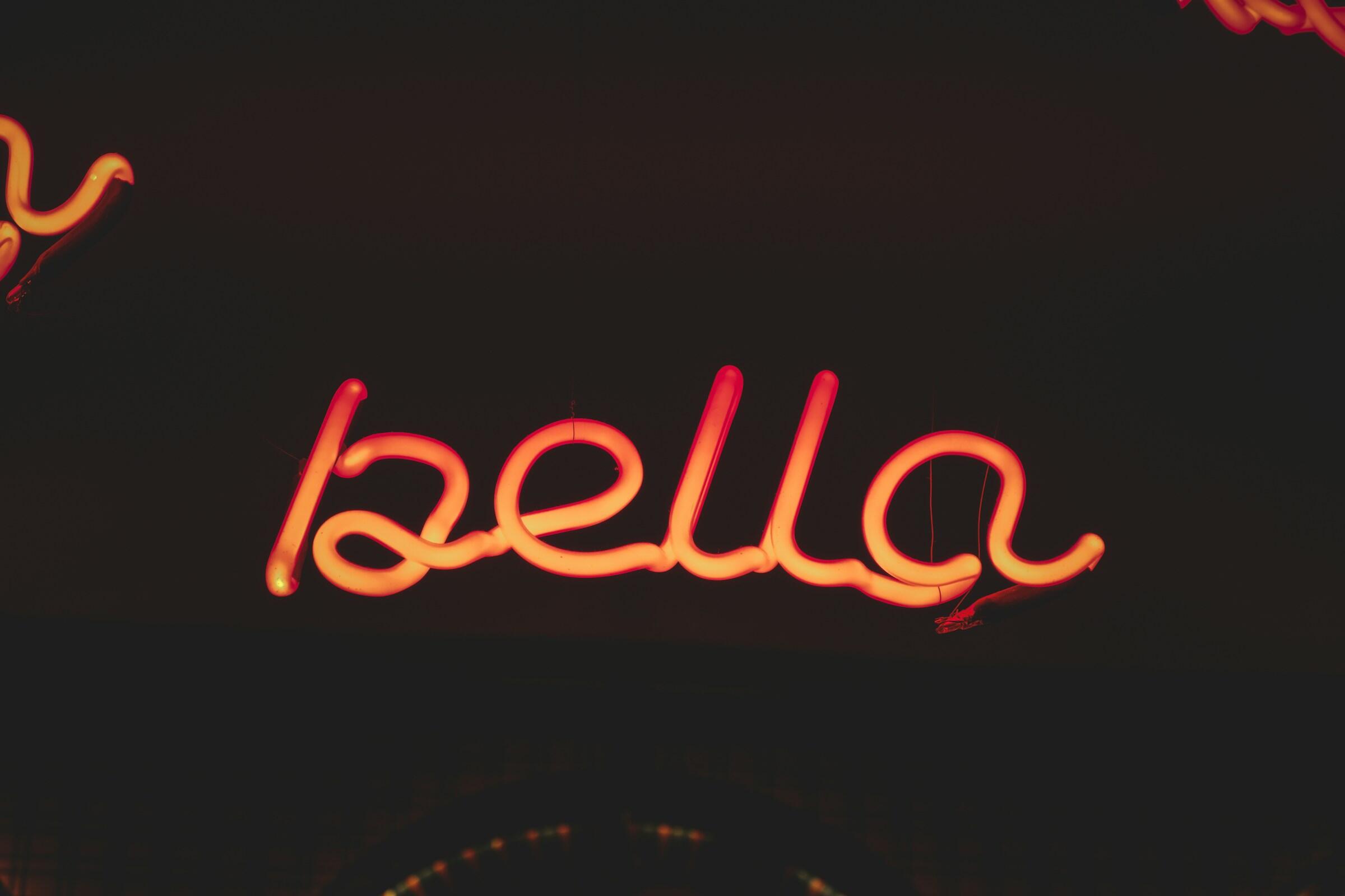 Un letrero luminoso de neón que muestra la palabra «bella» en elegante letra cursiva sobre un fondo oscuro.