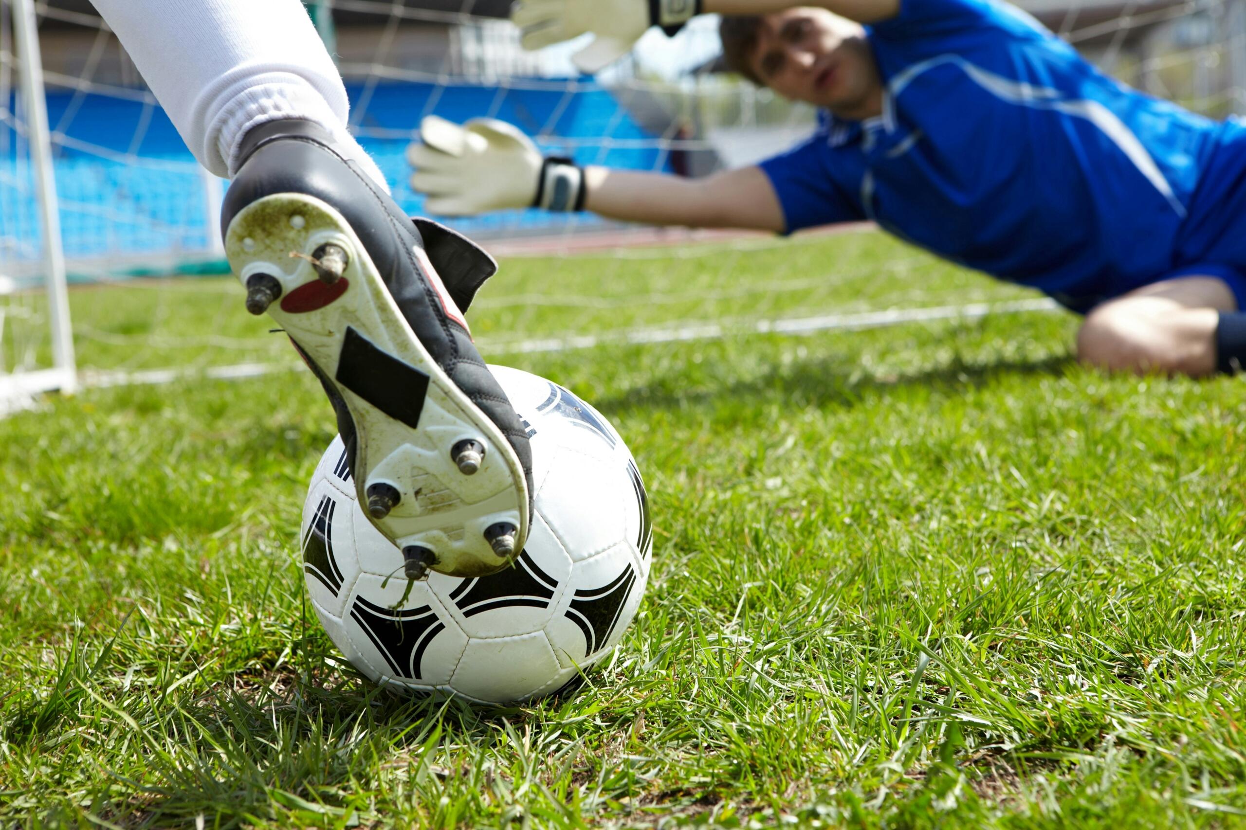 Un jugador de fútbol se prepara para chutar un balón en un campo de hierba, mientras un portero se estira para hacer una parada en el fondo.